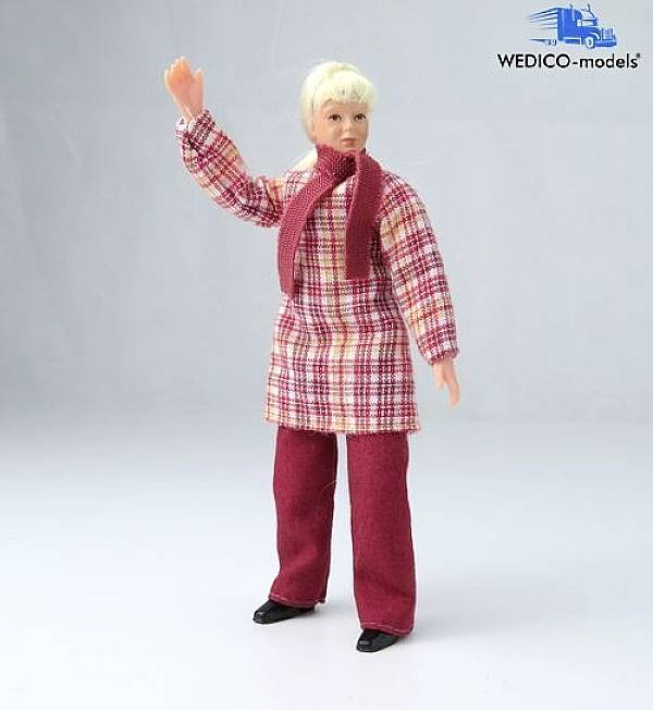 Lalka w kratowanym płaszczu i czerwonym szaliku unosi rękę, stojąc na jasnym tle.