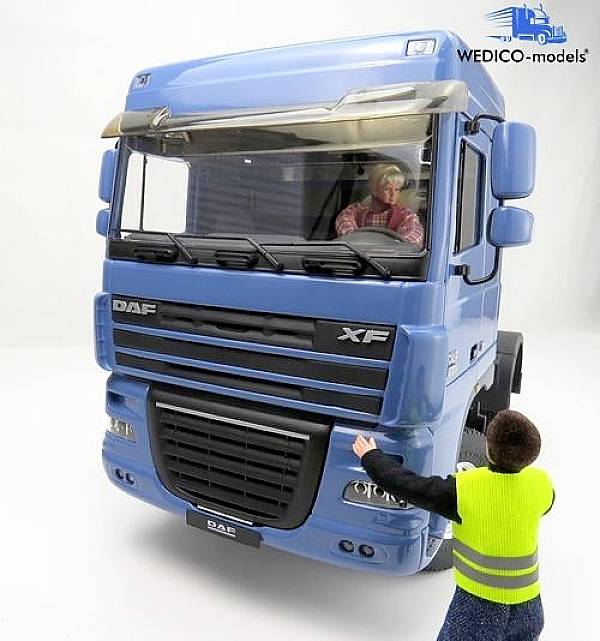 Duży niebieski samochód ciężarowy z kierowcą wewnątrz. Przed samochodem ciężarowym stoi osoba w żółtej kamizelce odblaskowej.