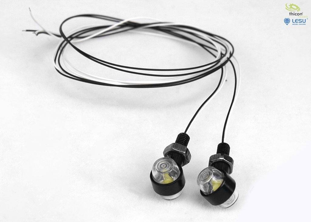 Dwie małe, okrągłe lampy LED z czarnymi przewodami i metalowymi zamknięciami, leżące na białym tle.