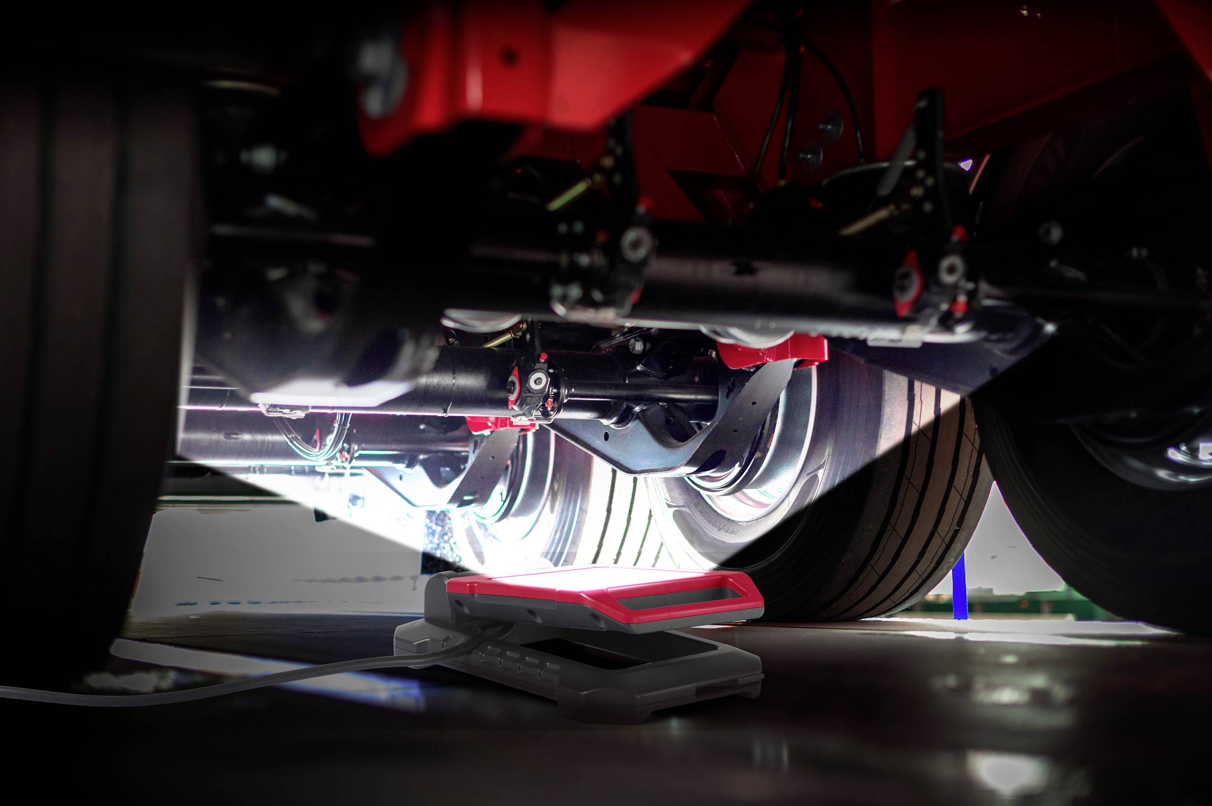 Pod czerwonym pojazdem znajduje się urządzenie techniczne do inspekcji lub pomiaru elementów zawieszenia.