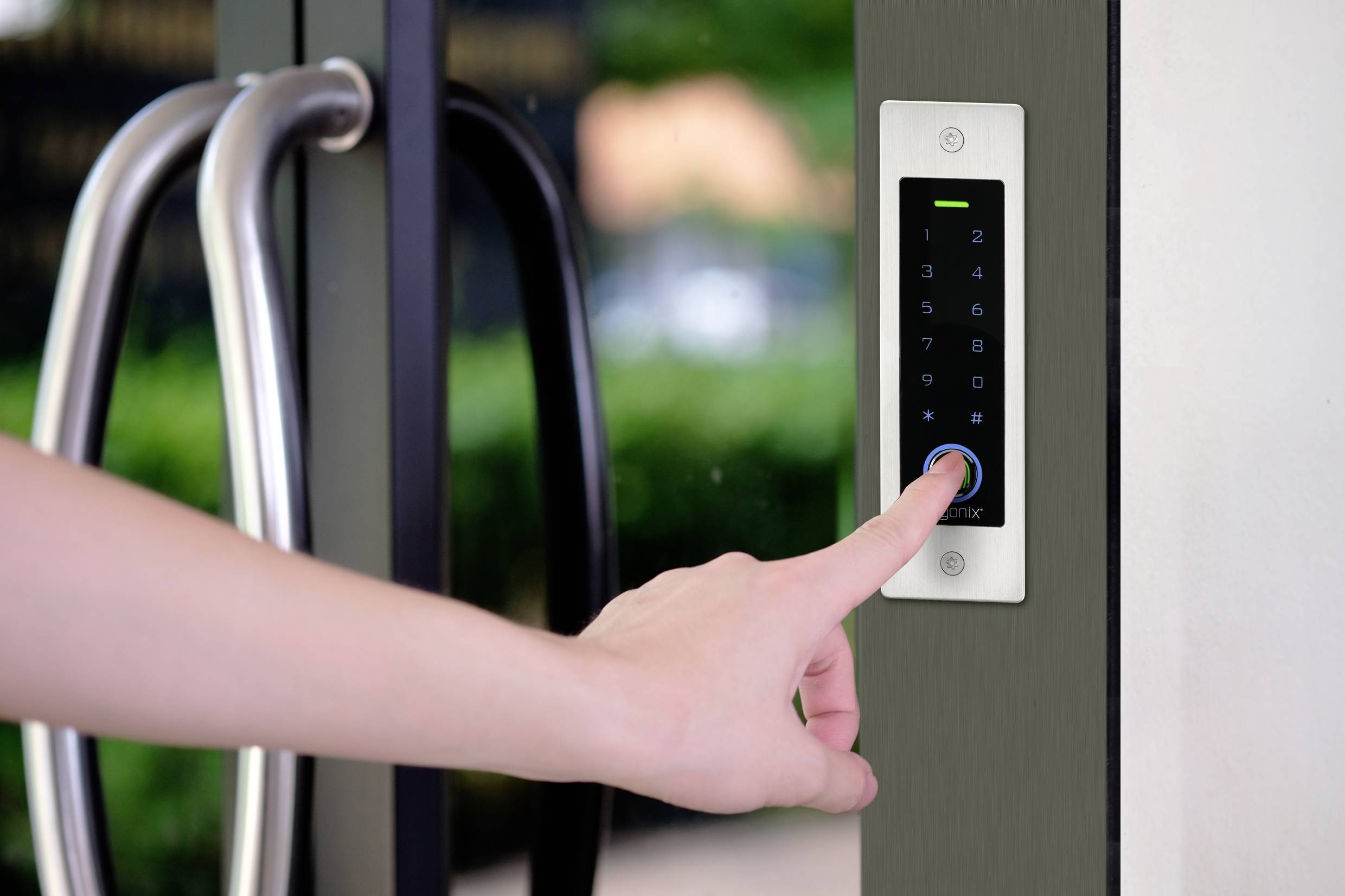 Ręka naciska ekran cyfrowego zamka przy szklanych drzwiach, wyświetlający klawiaturę numeryczną.