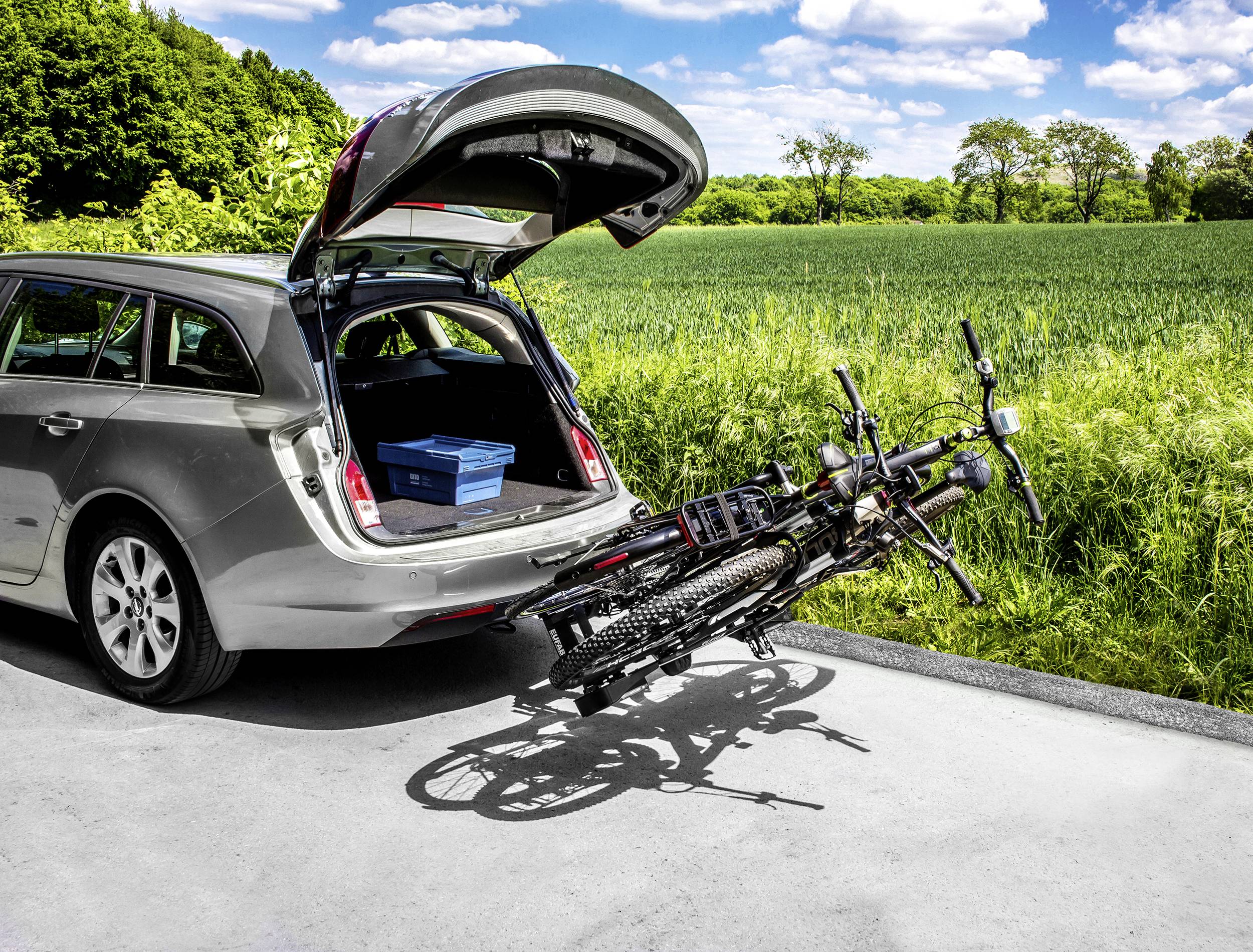 Srebrny samochód stoi przy krawędzi drogi z otwartym bagażnikiem, w którym znajduje się niebieskie pudło. Na samochodzie zamontowany jest bagażnik rowerowy z dwoma rowerami.