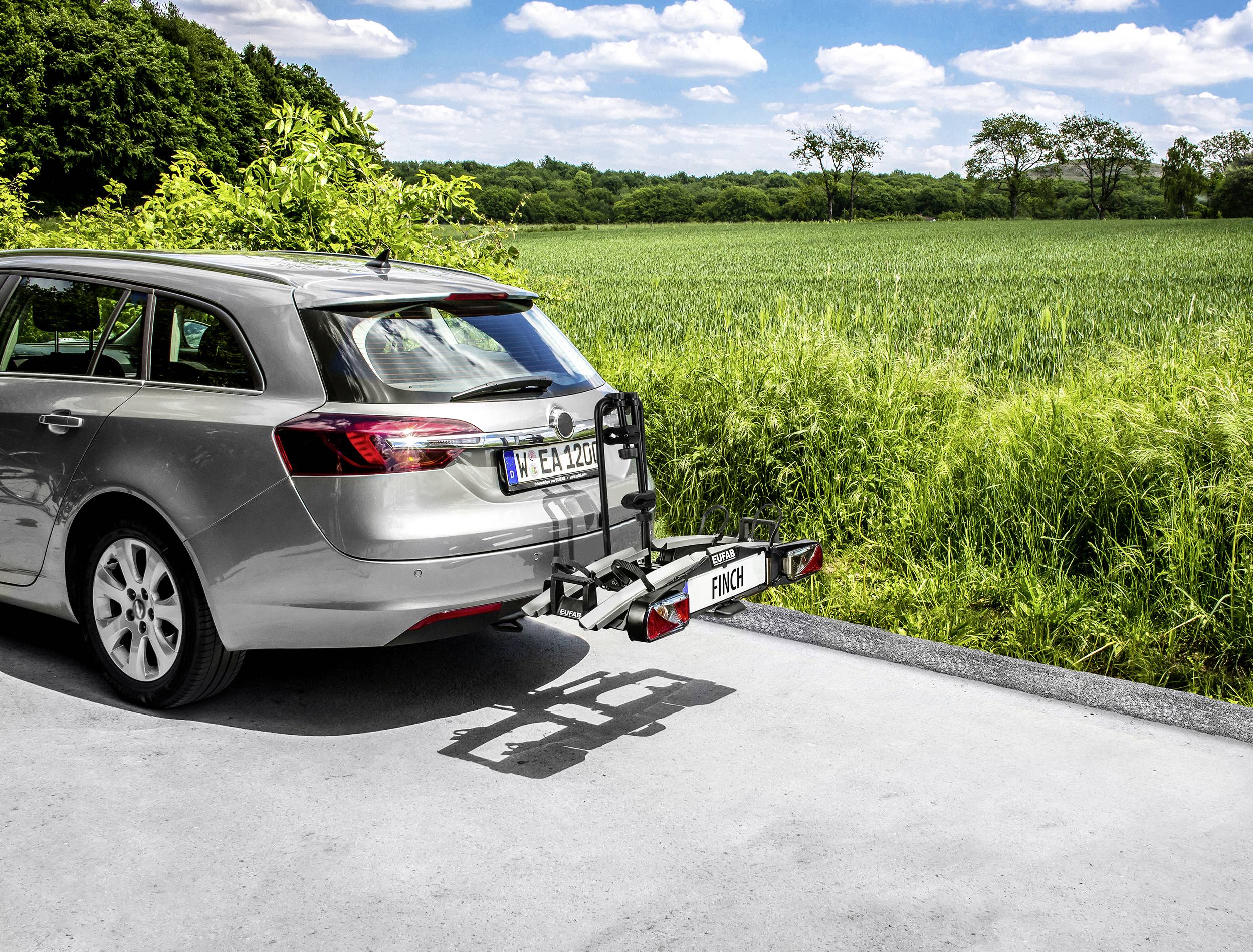 Srebrny samochód kombi z zamontowanym bagażnikiem rowerowym stoi na drodze obok zielonego pola pod błękitnym niebem z chmurami.