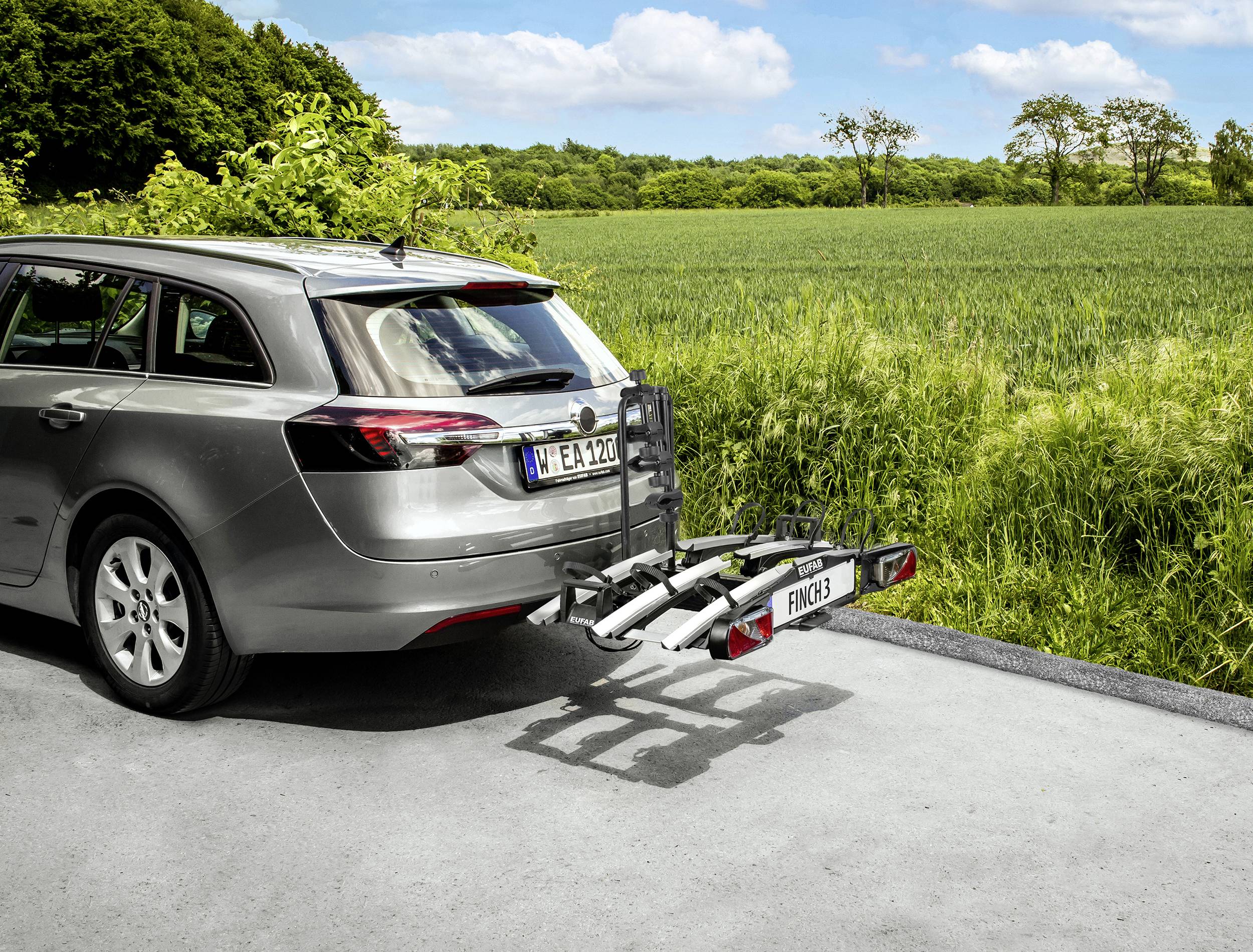 Samochód jedzie drogą z bagażnikiem rowerowym z tyłu, otoczony zielonymi polami i drzewami w tle.