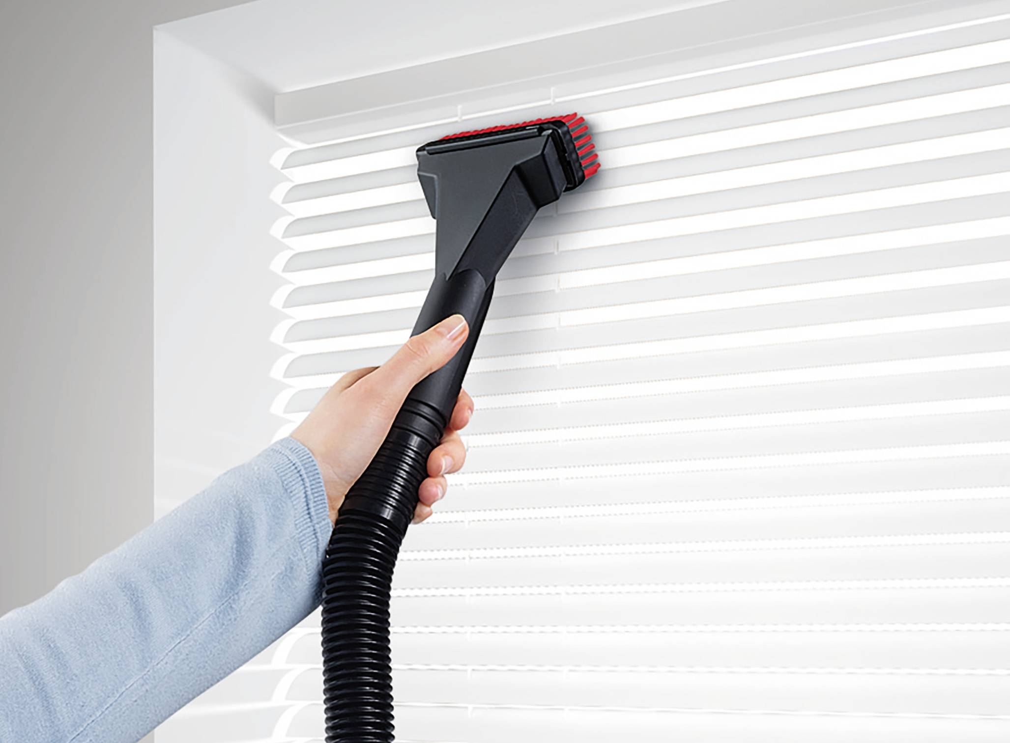 Osoba czyści białe żaluzje szczotką podłączoną do odkurzacza.