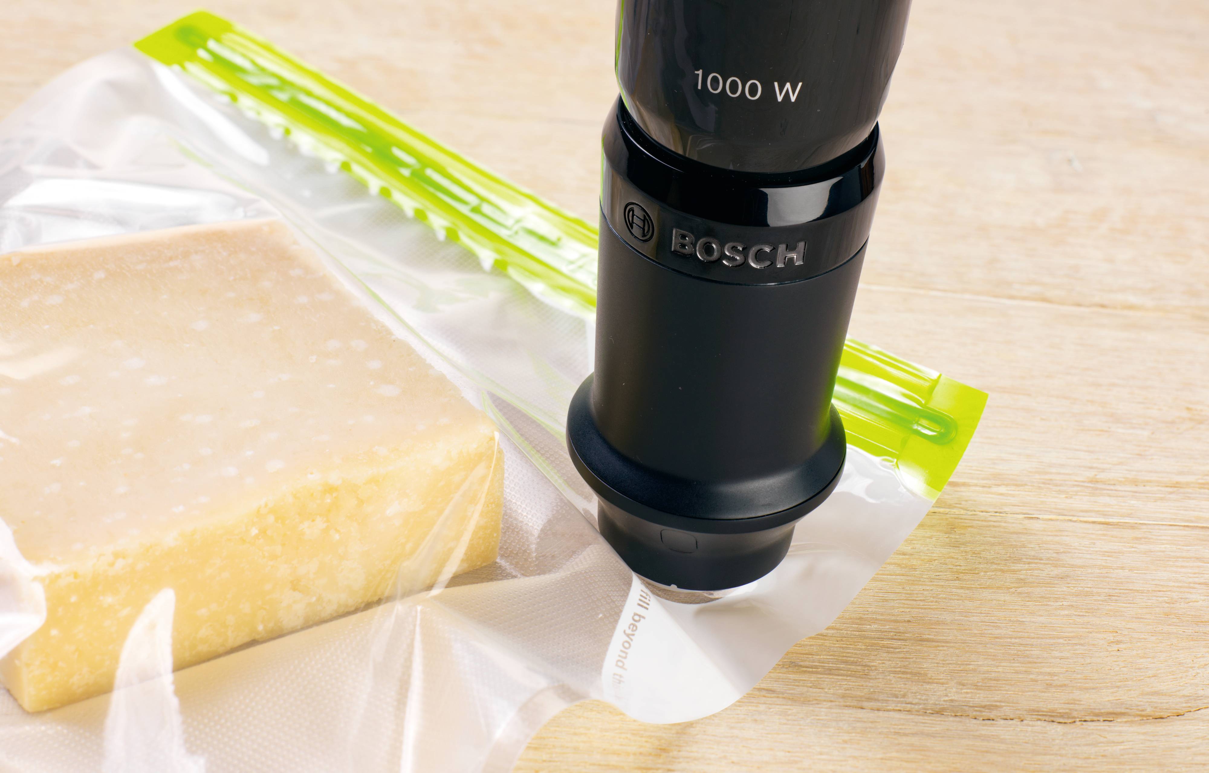 Maszyna próżniowa zamyka torbę z kawałkiem sera. Urządzenie oznaczone '1000 W'. Drewniany stół w tle.