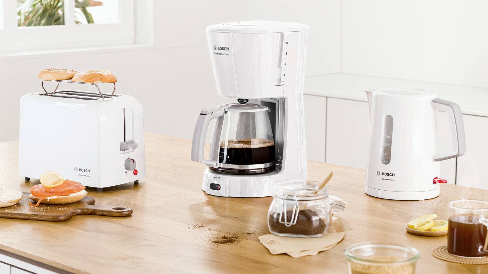 Scena kuchenna z tosterem, ekspresem do kawy i czajnikiem elektrycznym na stole. Widoczny jest również jeden plaster tosta, kawa oraz dżem.