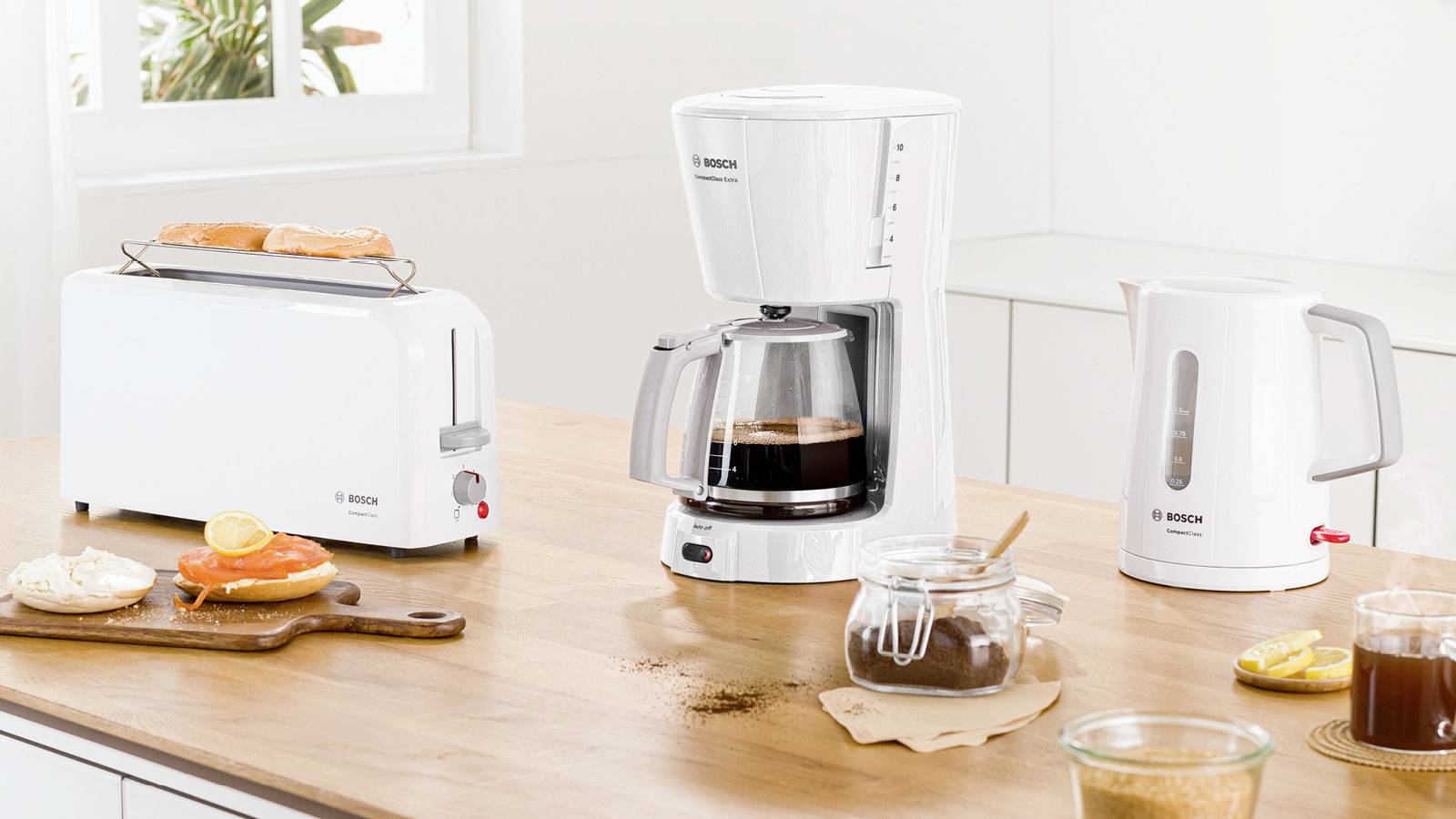 Biały toster na drewnianym blacie wypieka chleb. Obok niego stoi ekspres do kawy z pełnym dzbankiem i czajnik elektryczny.