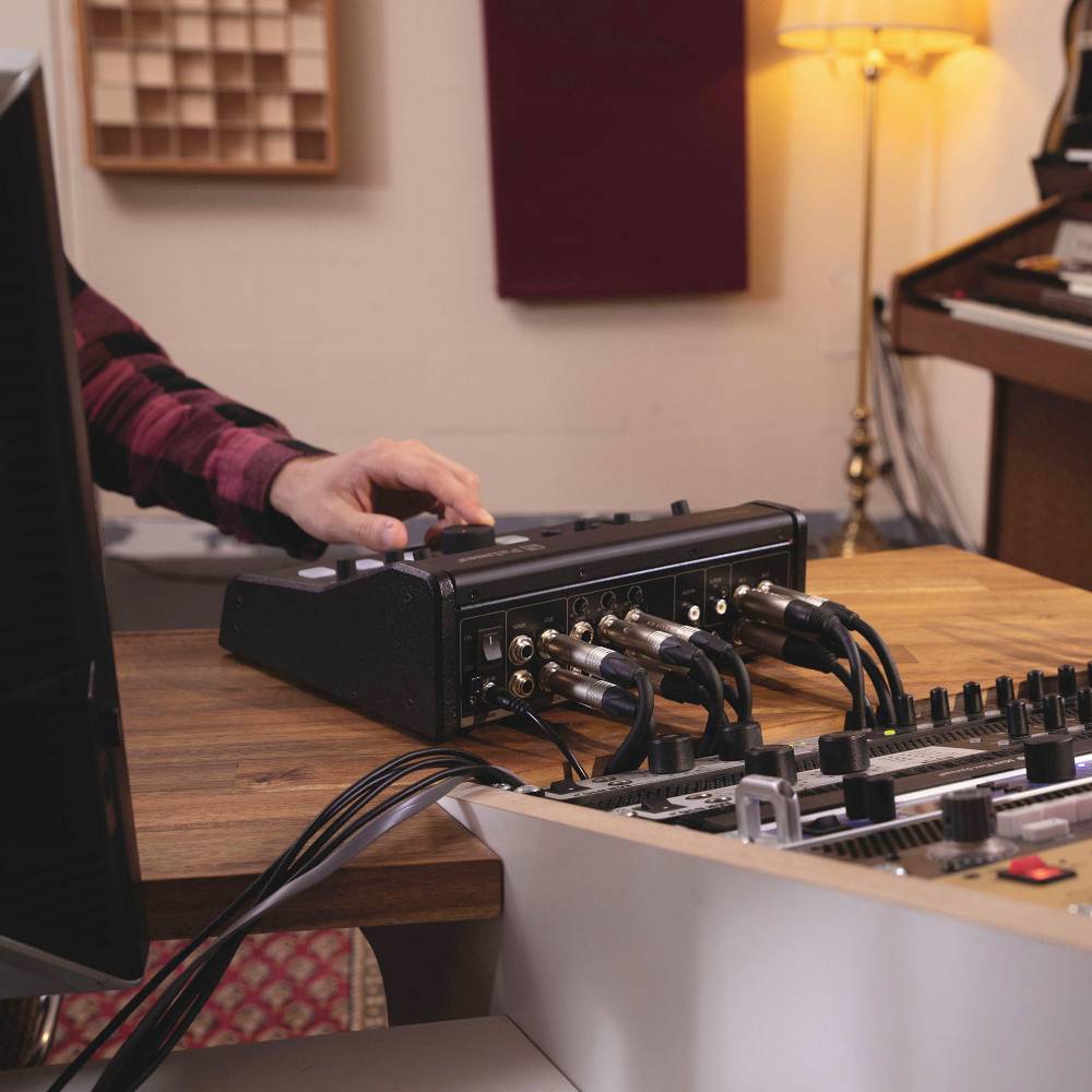 Nagranie w studio nagrań: Osoba reguluje urządzenia audio przy stole, otoczona konsoletą mikserską i kablami. W tle lampa i dekoracja ścienna.