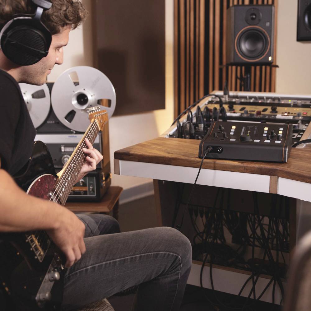 Muzyk w studio nagraniowym gra na gitarze elektrycznej, nosi słuchawki. W tle magnetofony i mikser.