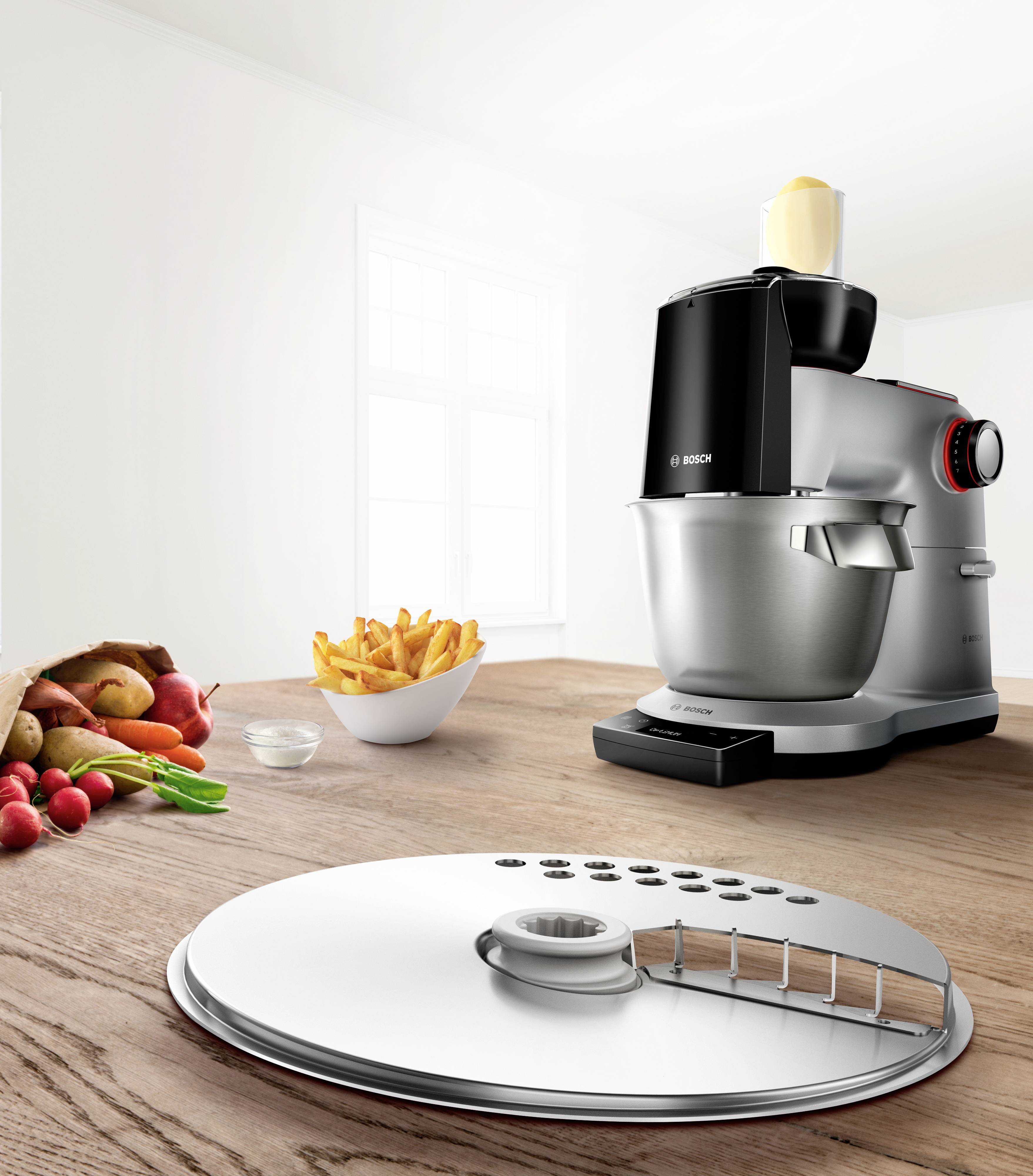 Robot kuchenny na drewnianym blacie, obok niego nóż do ciasta i miska frytek; świeże warzywa w tle; nowoczesna, jasna kuchnia.