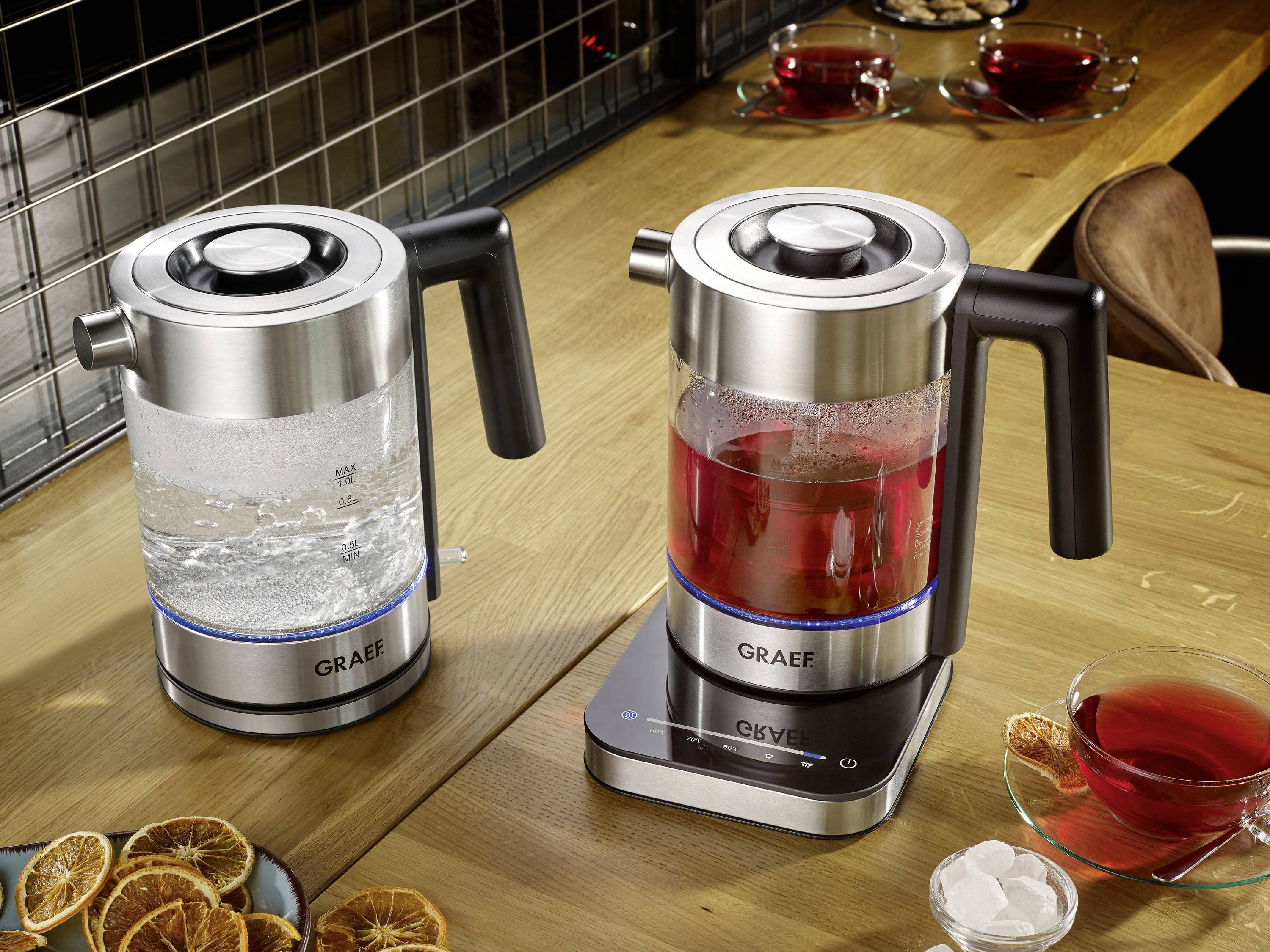 Dwa szklane czajniki na blacie kuchennym: po lewej stronie wrząca woda, po prawej czerwona herbata. Obok szklana taca z przezroczystą filiżanką.