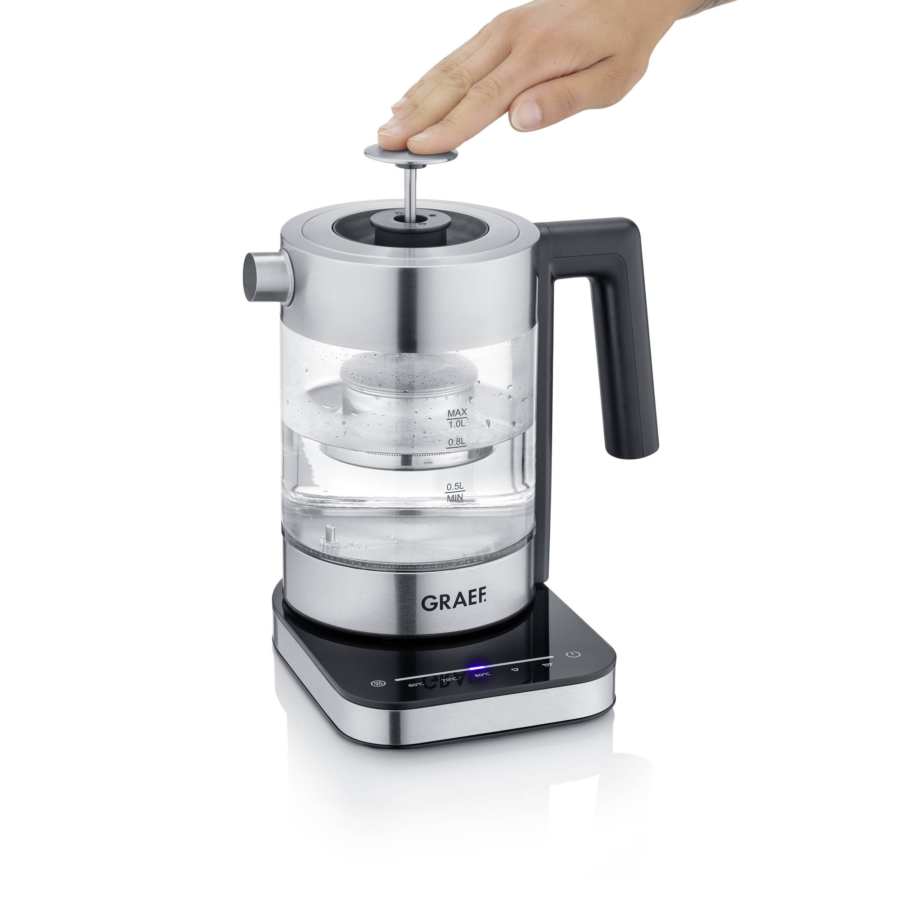 Elektryczny czajnik z przezroczystego szkła z czarnym uchwytem. Ręka jest w trakcie otwierania pokrywy. Z przodu widoczna jest marka 'Graef'.