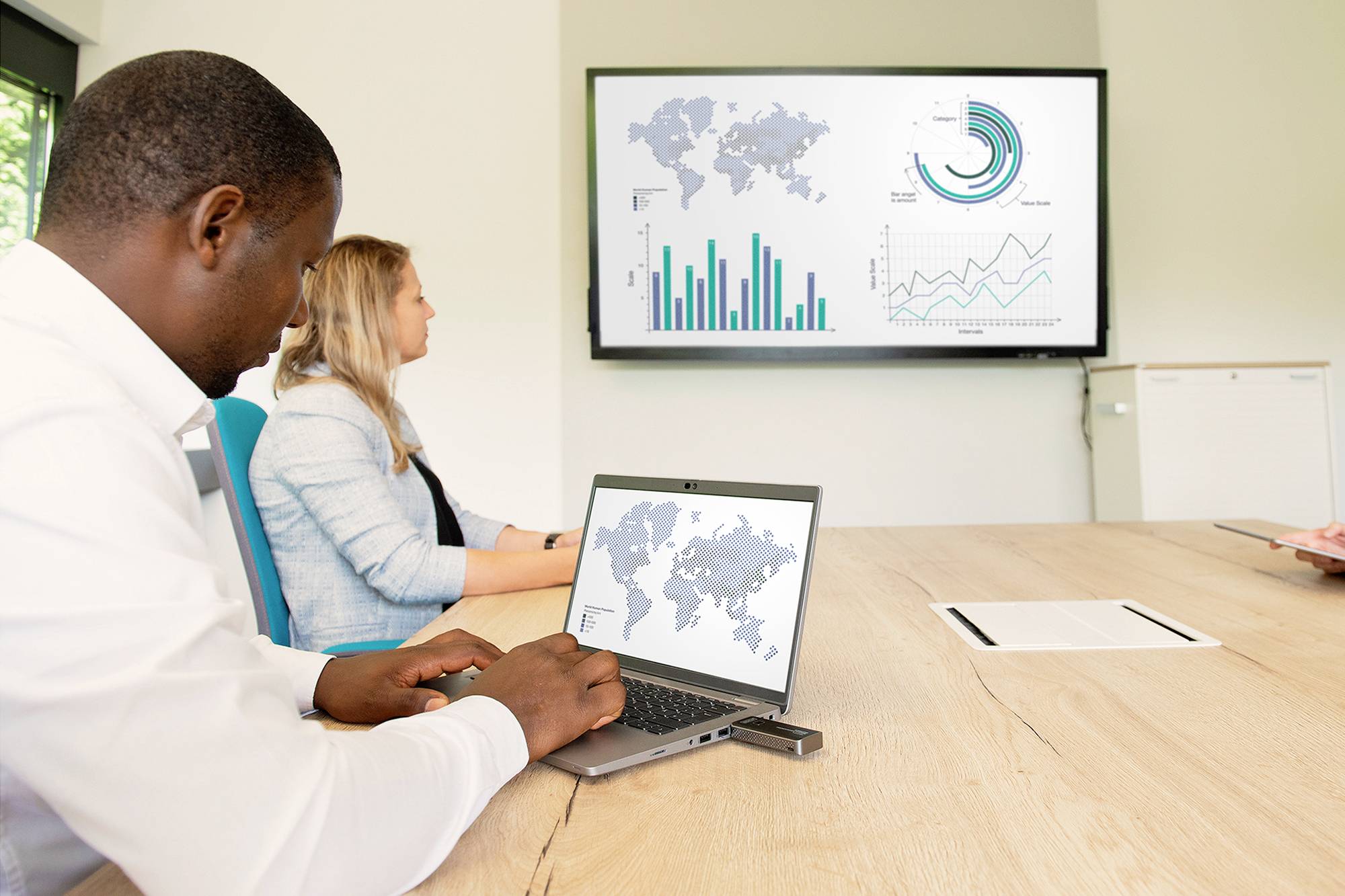 Dwie osoby w sali konferencyjnej przyglądają się ekranowi z grafikami i diagramami przedstawiającymi dane globalne. Jedna z osób pracuje na laptopie.