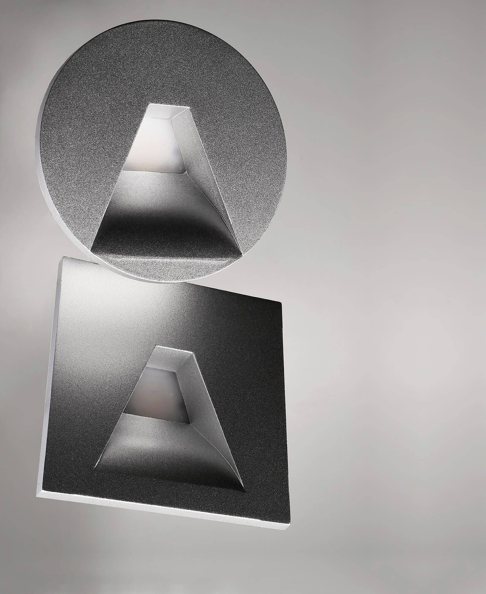Srebrzyste, geometryczne lampy na szarej ścianie, jedna okrągła, druga kwadratowa, każda z trójkątnym otworem świetlnym.