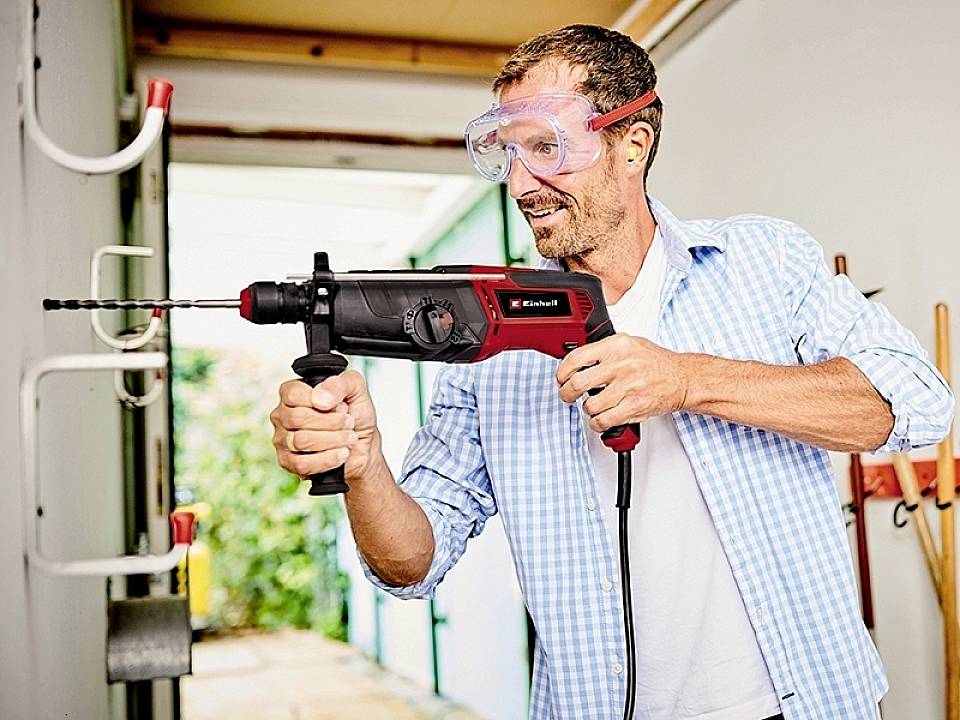 Mężczyzna wierca dziurę w ścianie za pomocą wiertarki. Ma na sobie okulary ochronne i uśmiecha się podczas wykonywania zadania.