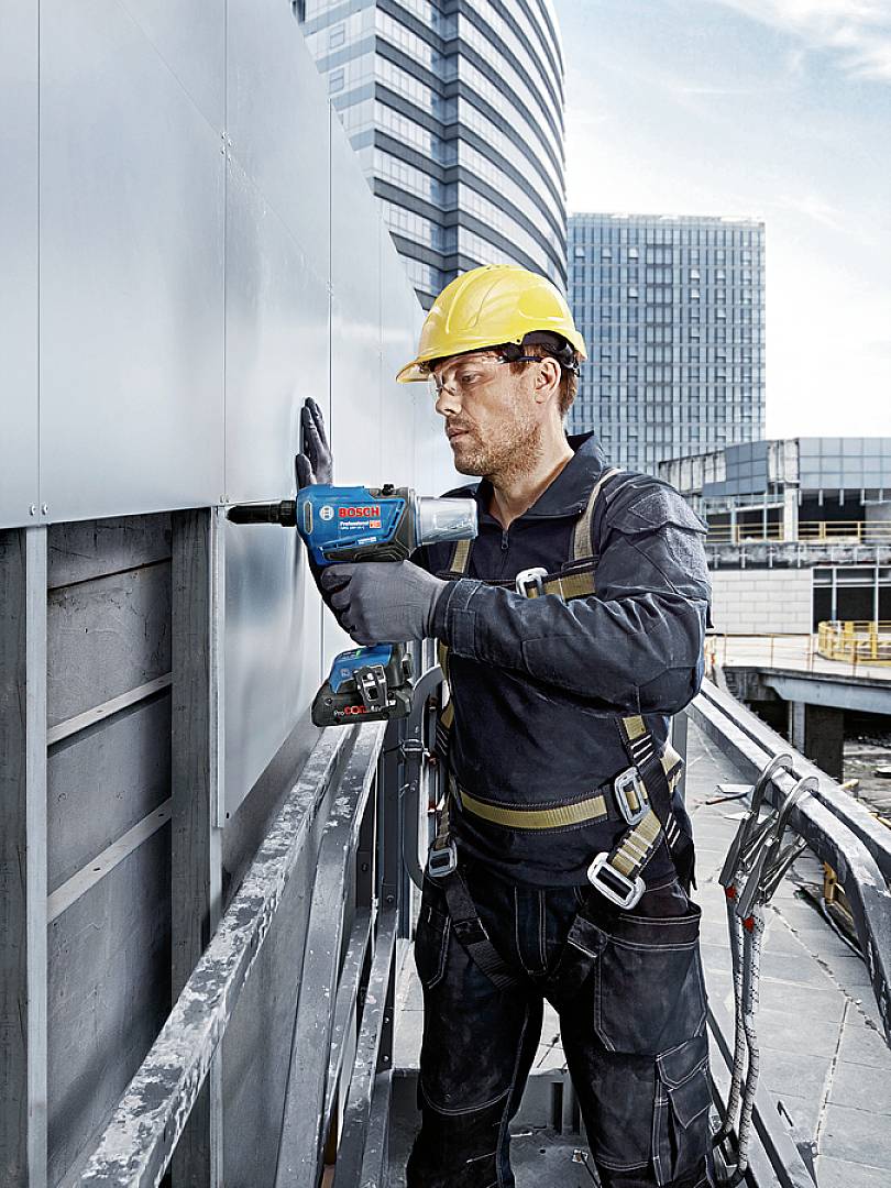 Robotnik budowlany w kasku i uprzęży bezpieczeństwa wiertający otwór w metalowej ścianie, na tle nowoczesnych budynków placu budowy.