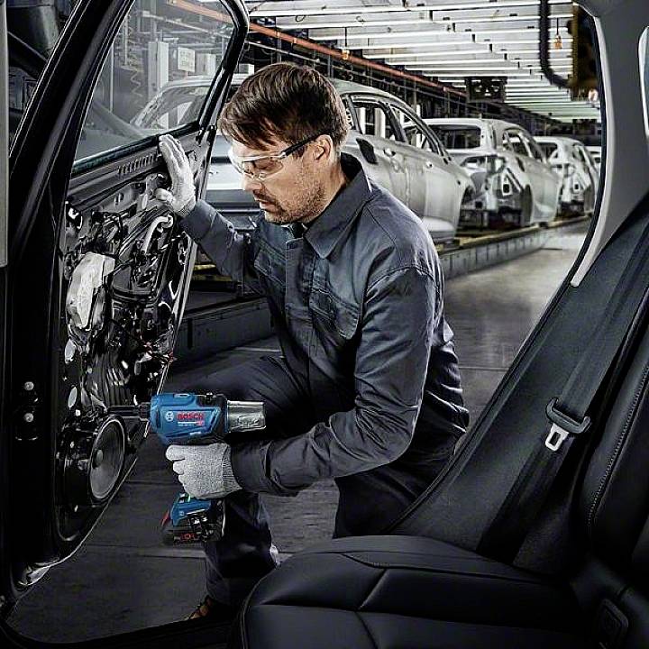 Technik montuje części drzwi pojazdu za pomocą elektronarzędzia w fabryce samochodów. W tle widać kilka samochodów na linii produkcyjnej.