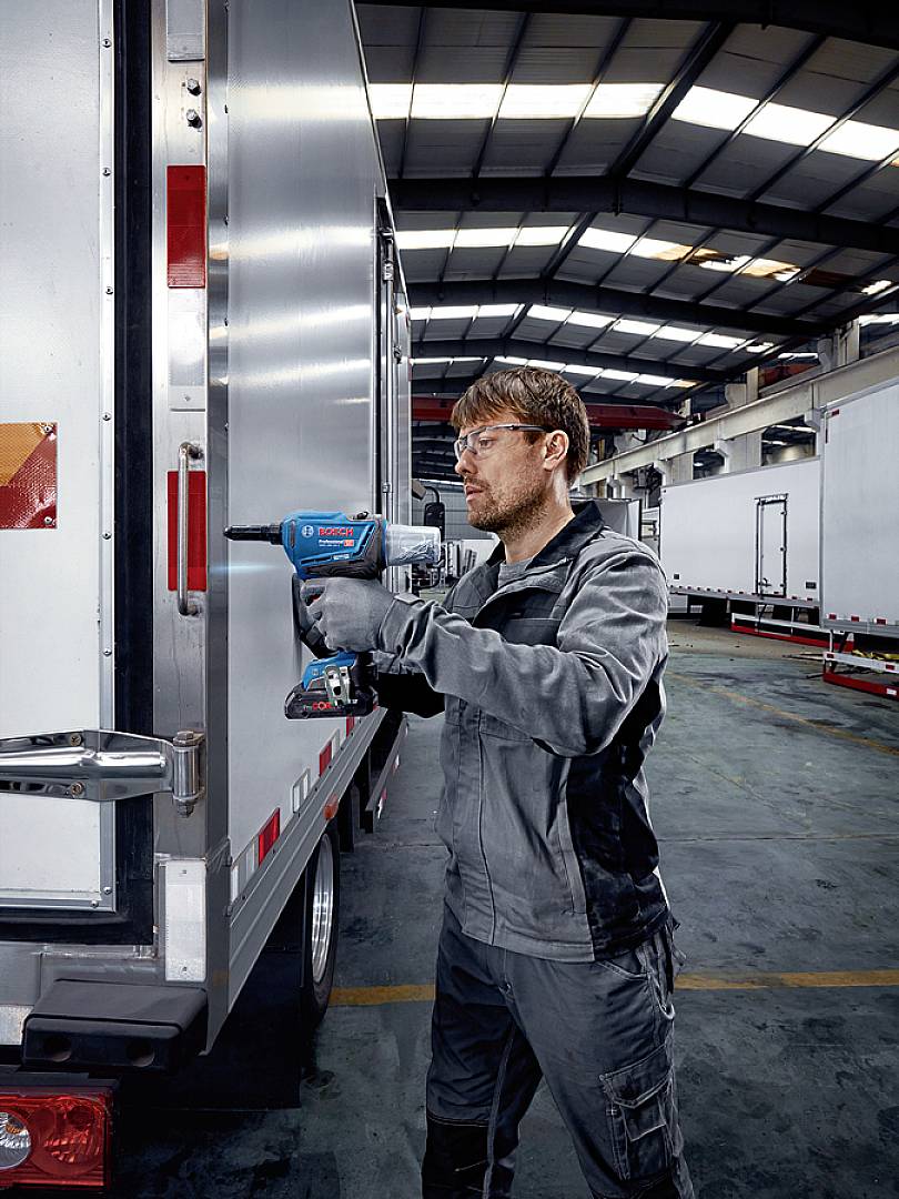 Osoba w ubraniu roboczym używa akumulatorowego wkrętarki do przeprowadzania napraw na ciężarówce w dużej hali magazynowej.