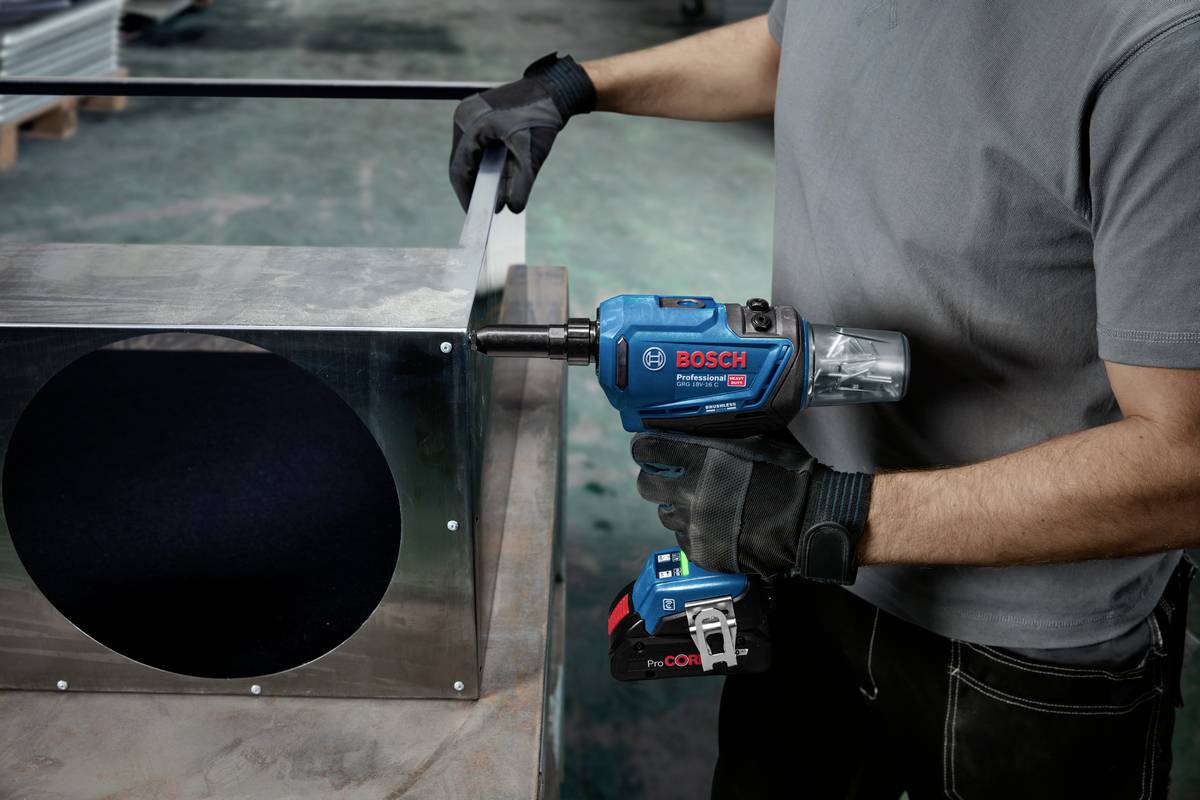 Osoba obrabia metal niebieską wiertarką. Wiertło jest używane przy dużym metalowym pudełku. Pracownik nosi czarne rękawice.