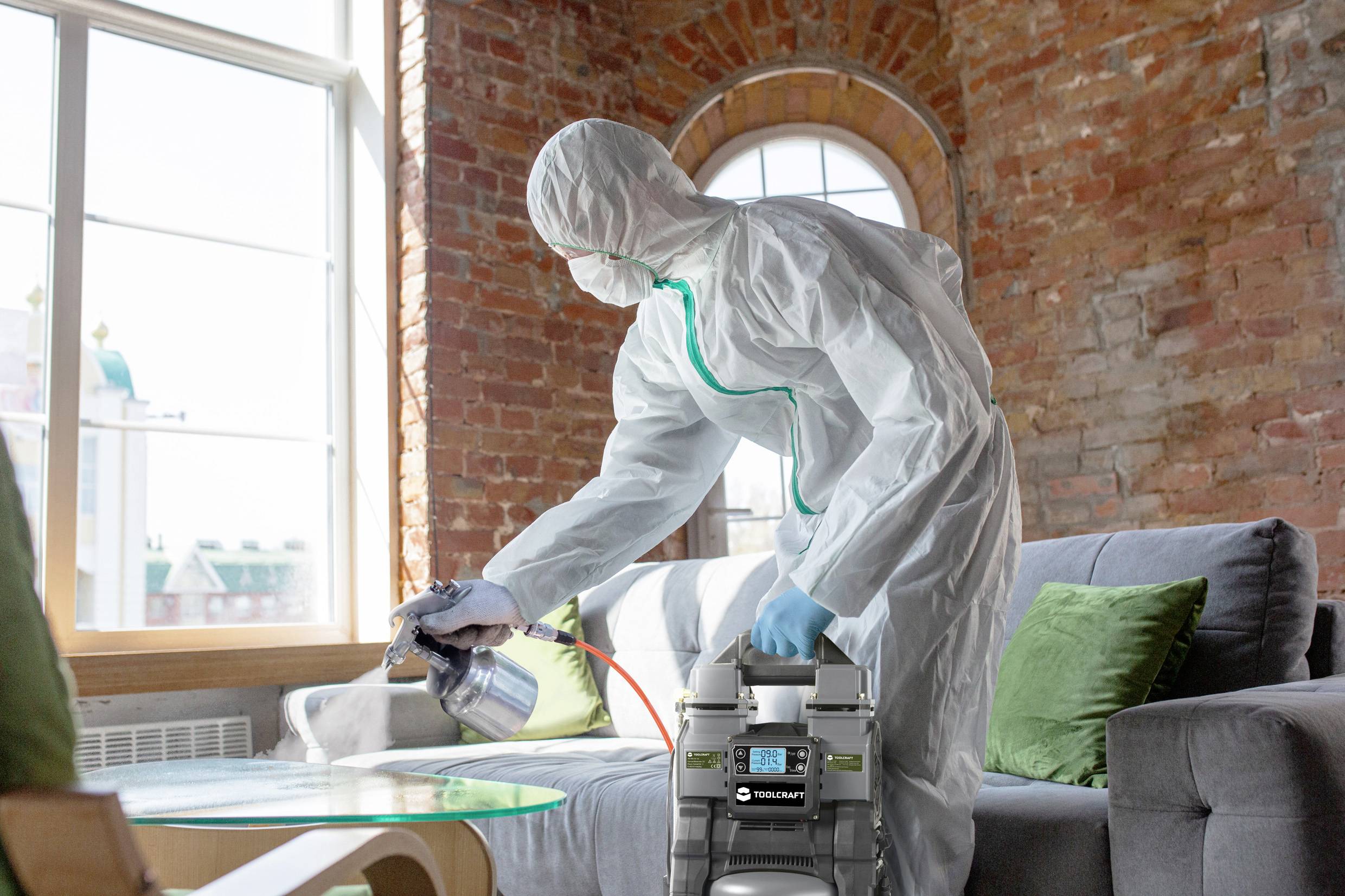 Osoba w kombinezonie ochronnym dezynfekuje pomieszczenie mieszkalne za pomocą urządzenia natryskowego. Tablica wisząca na ceglastej ścianie i sofa w tle.