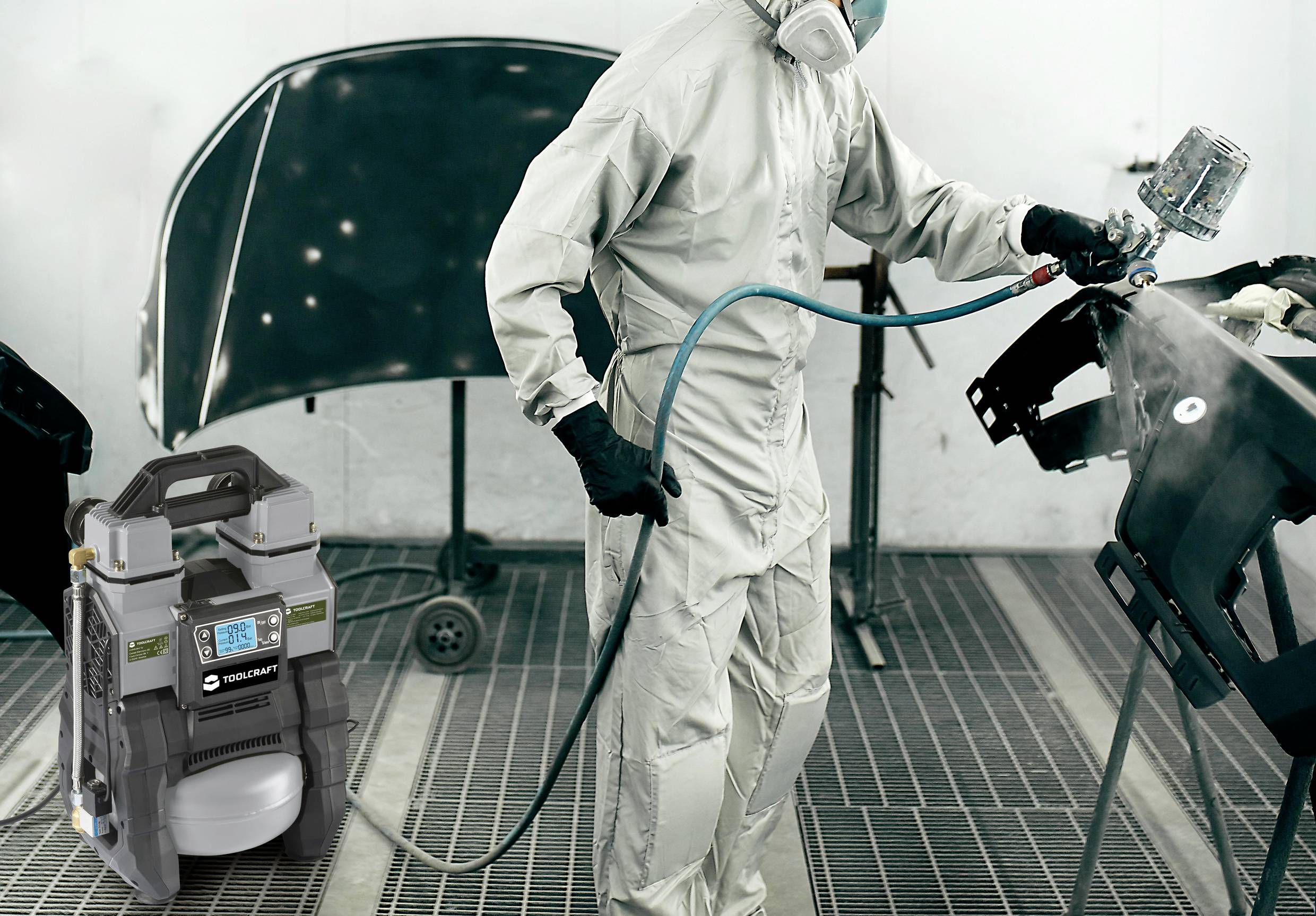 Osoba w kombinezonie ochronnym maluje części samochodowe w warsztacie. Na pierwszym planie stoi przenośny kompresor.