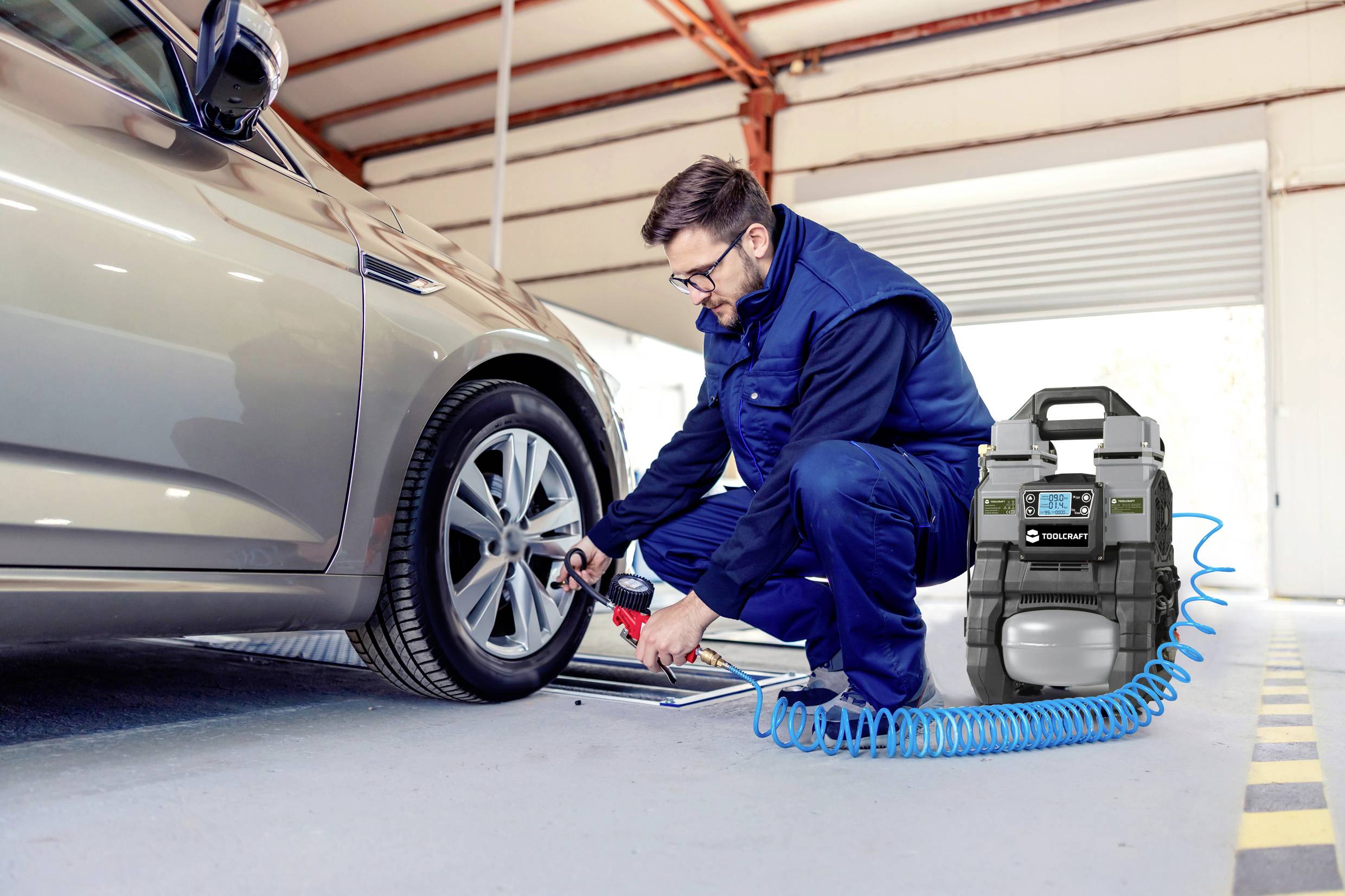 Mechanik sprawdza ciśnienie powietrza w oponach samochodu osobowego w warsztacie. Obok niego stoi sprężarka powietrza z podłączonym wężem.