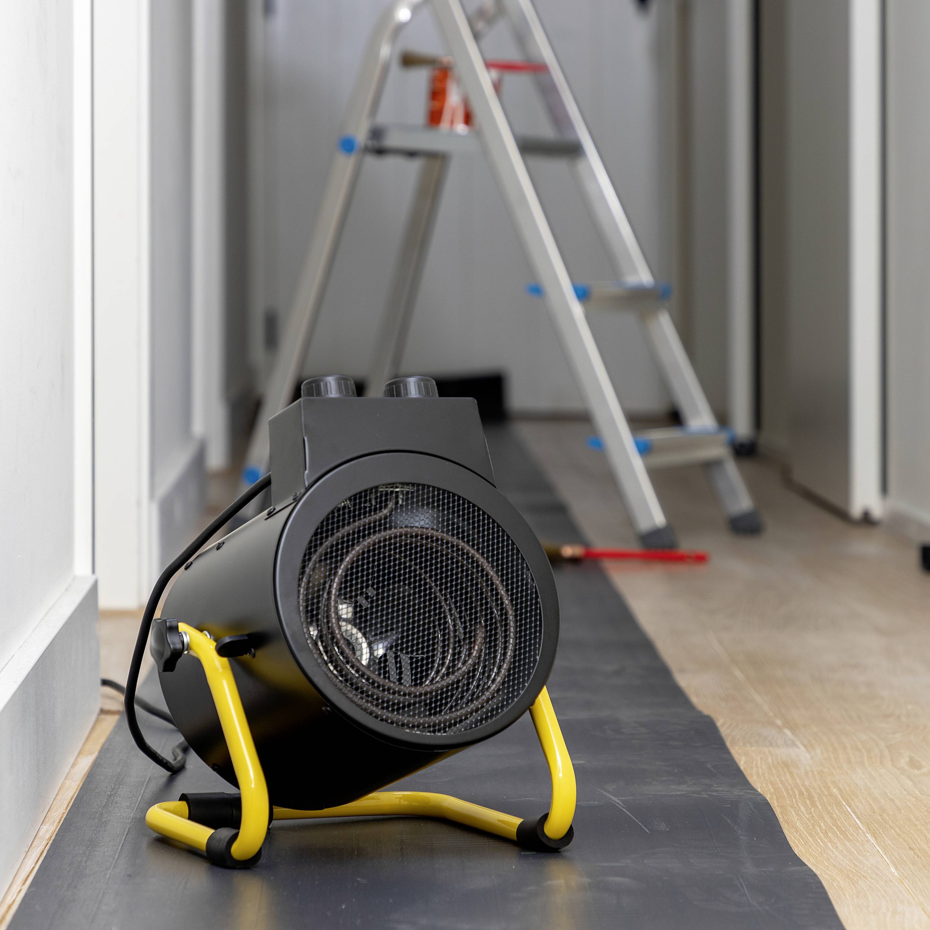 Grzejnik elektryczny stojący na podłodze korytarza, w tle wysuwana drabina. Scena sugeruje trwające prace remontowe.