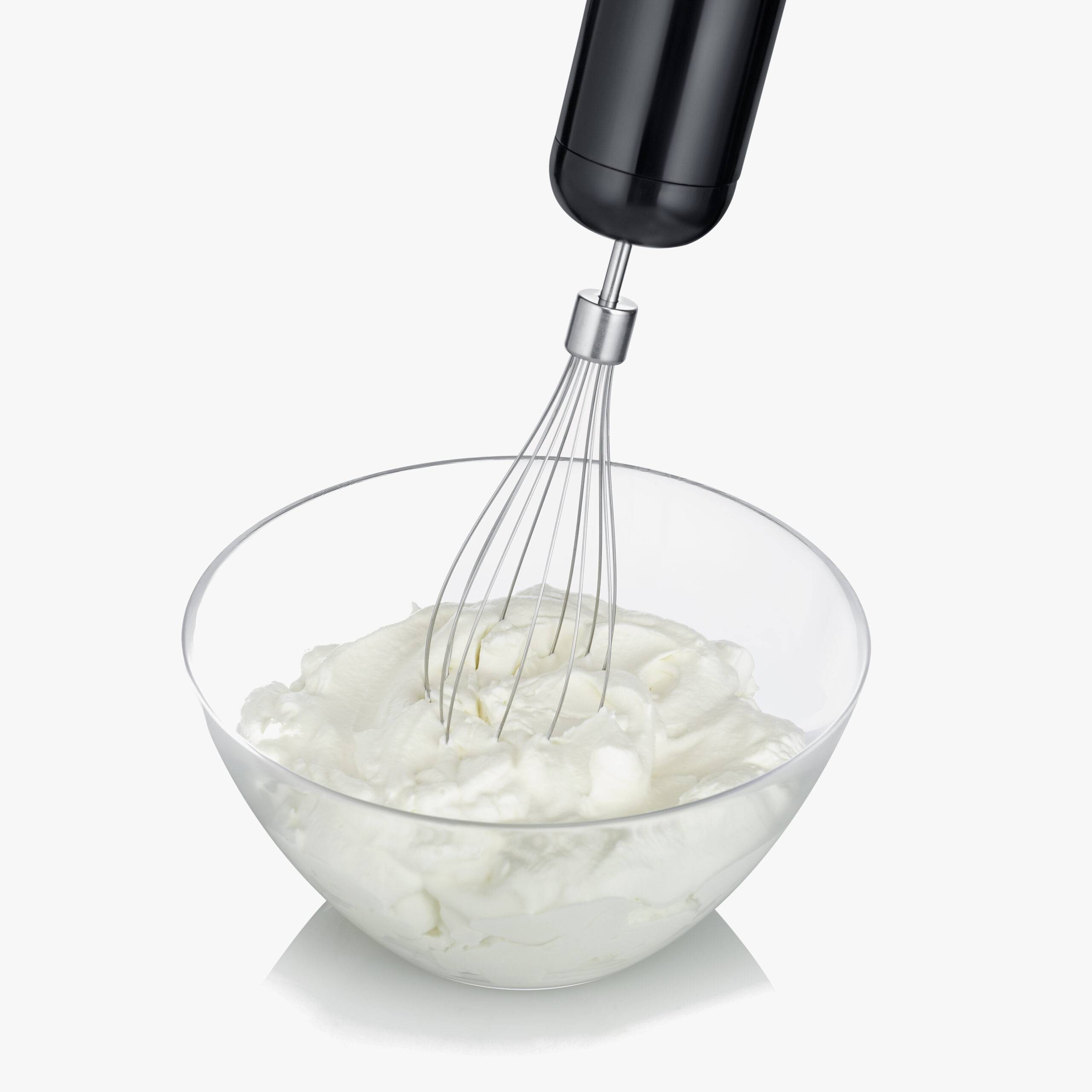 Elektryczny mikser ręczny ubija śmietanę w przezroczystej misce. Śmietana zaczyna gęstnieć.