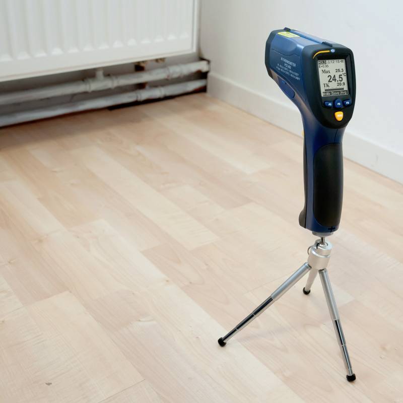 Miernik temperatury podczerwieni ze statywem stoi na drewnianej podłodze obok grzejnika. Wyświetlacz wskazuje 24,5°C.