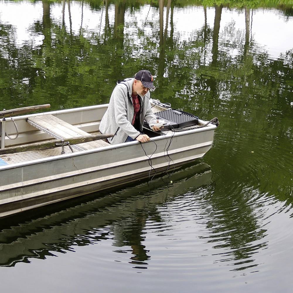 Mężczyzna siedzi w małej łodzi na spokojnym jeziorze i pracuje na urządzeniach elektronicznych, otoczony drzewami na brzegu.