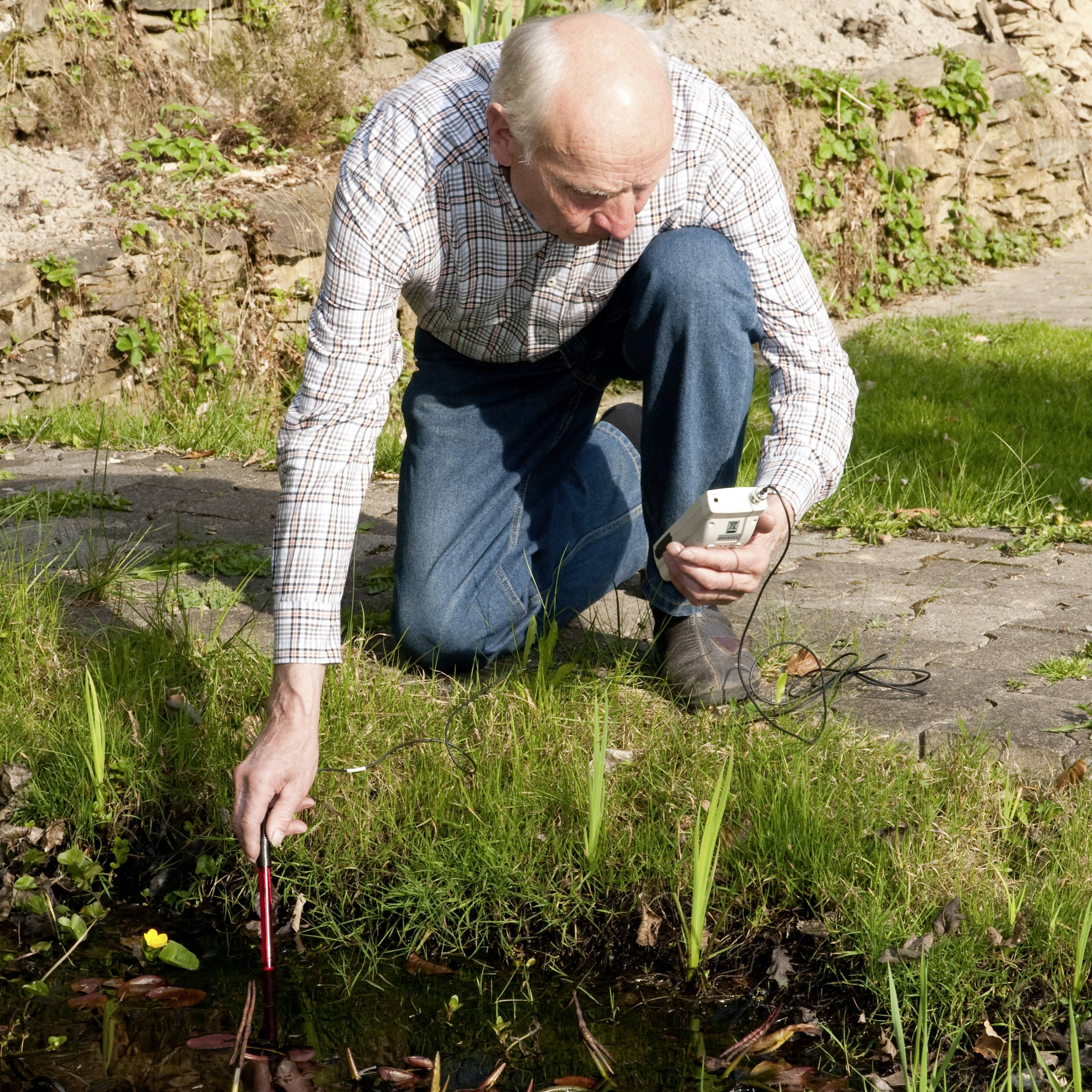 Osoba bada rośliny nad stawem za pomocą przyrządu pomiarowego, otoczona kamieniami i trawą.