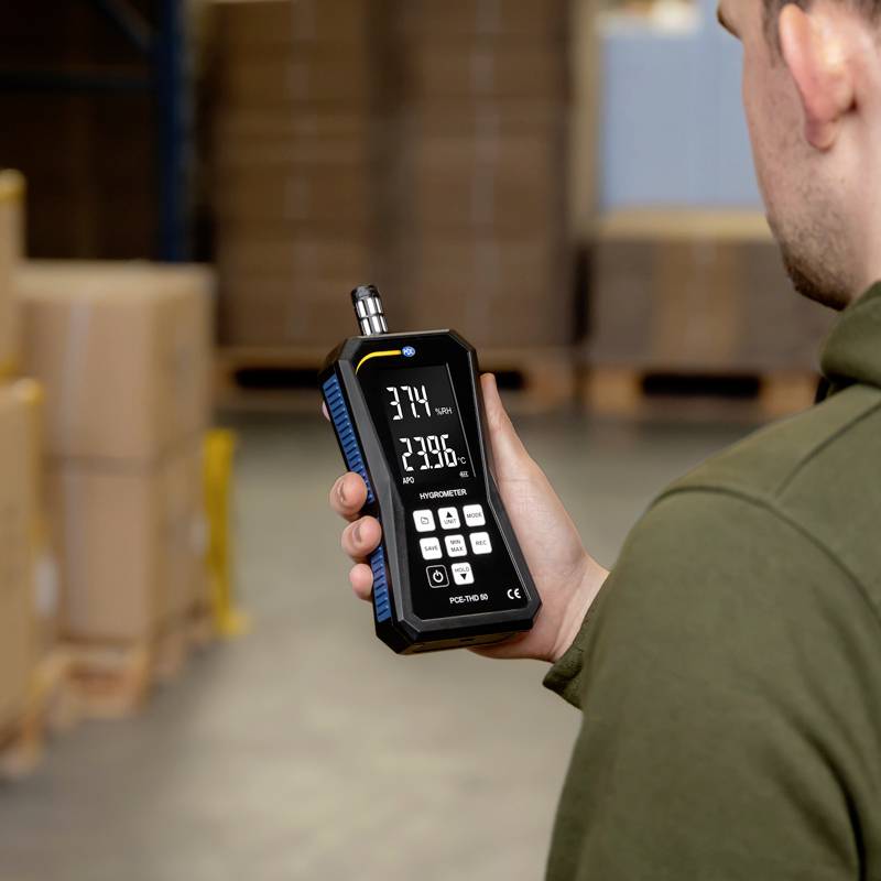 Mężczyzna trzyma ręczne urządzenie pomiarowe w magazynie. Na wyświetlaczu widoczne są wartości pomiarowe, podczas gdy stoi przed ułożonymi w stos kartonami.