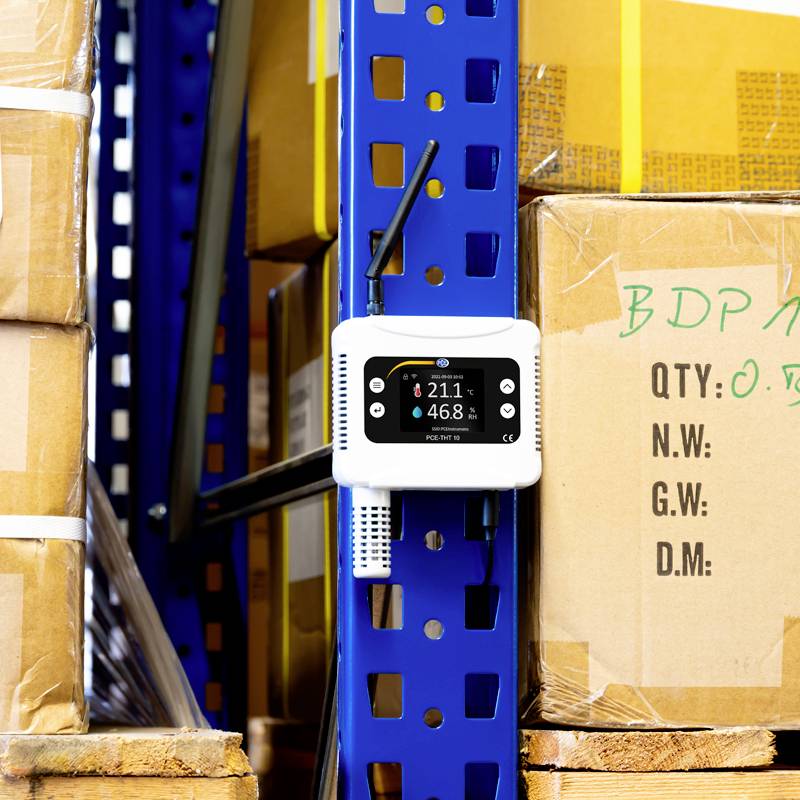 Czujnik temperatury i wilgotności wisi na niebieskim regale w magazynie. Obok niego są ułożone w stos brązowe kartony.