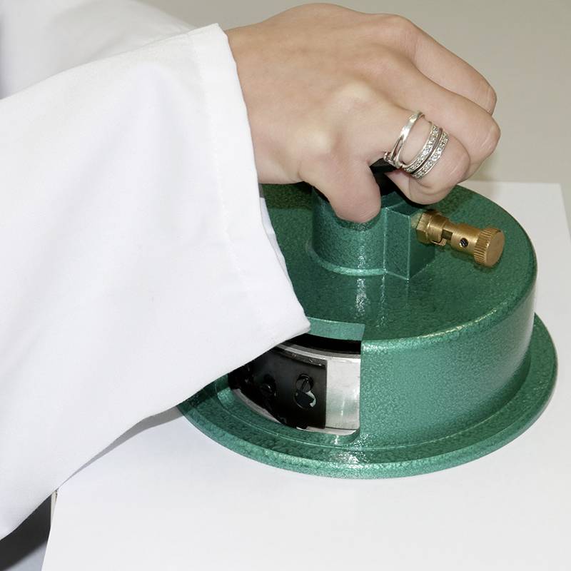 Dłoń w białym kitlu laboratoryjnym trzyma zielone urządzenie laboratoryjne. Osoba ma na palcach kilka pierścieni.