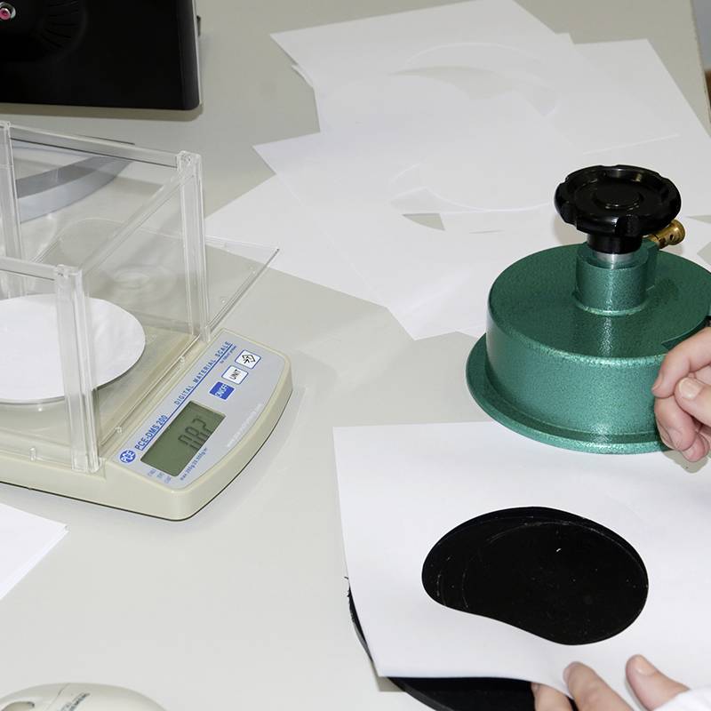 Osoba trzyma białą kartkę papieru z czarnym kołem. Po prawej stronie znajduje się zielone urządzenie. Po lewej stronie elektroniczna waga.