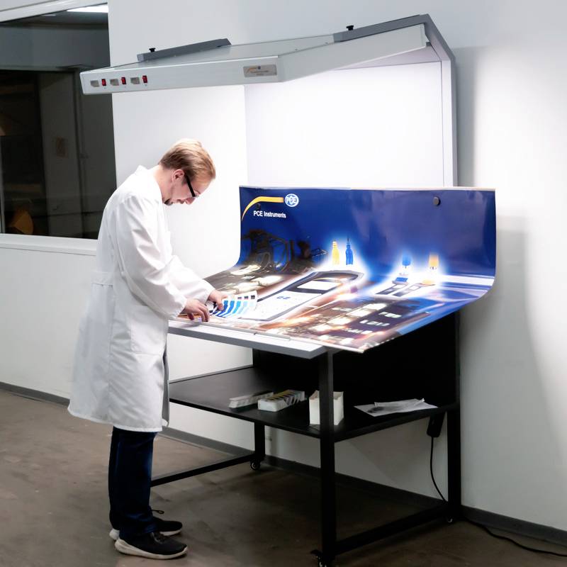 Mężczyzna w białym kitlu laboratoryjnym stoi przed stołem świetlnym w laboratorium, analizując próbki materiałów w znormalizowanym oświetleniu.