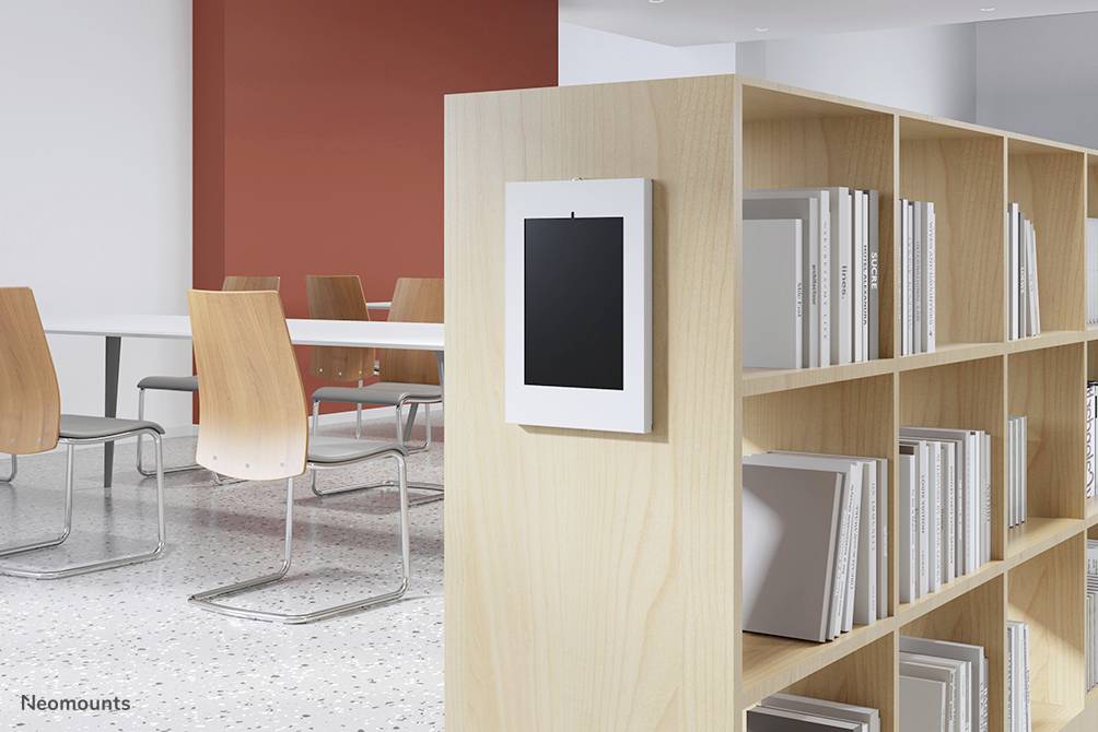 Nowoczesna przestrzeń biblioteczna z regałem na książki, drewnianymi krzesłami i czerwonym akcentem na ścianie. Tablet jest zamocowany przy jednym z regałów.