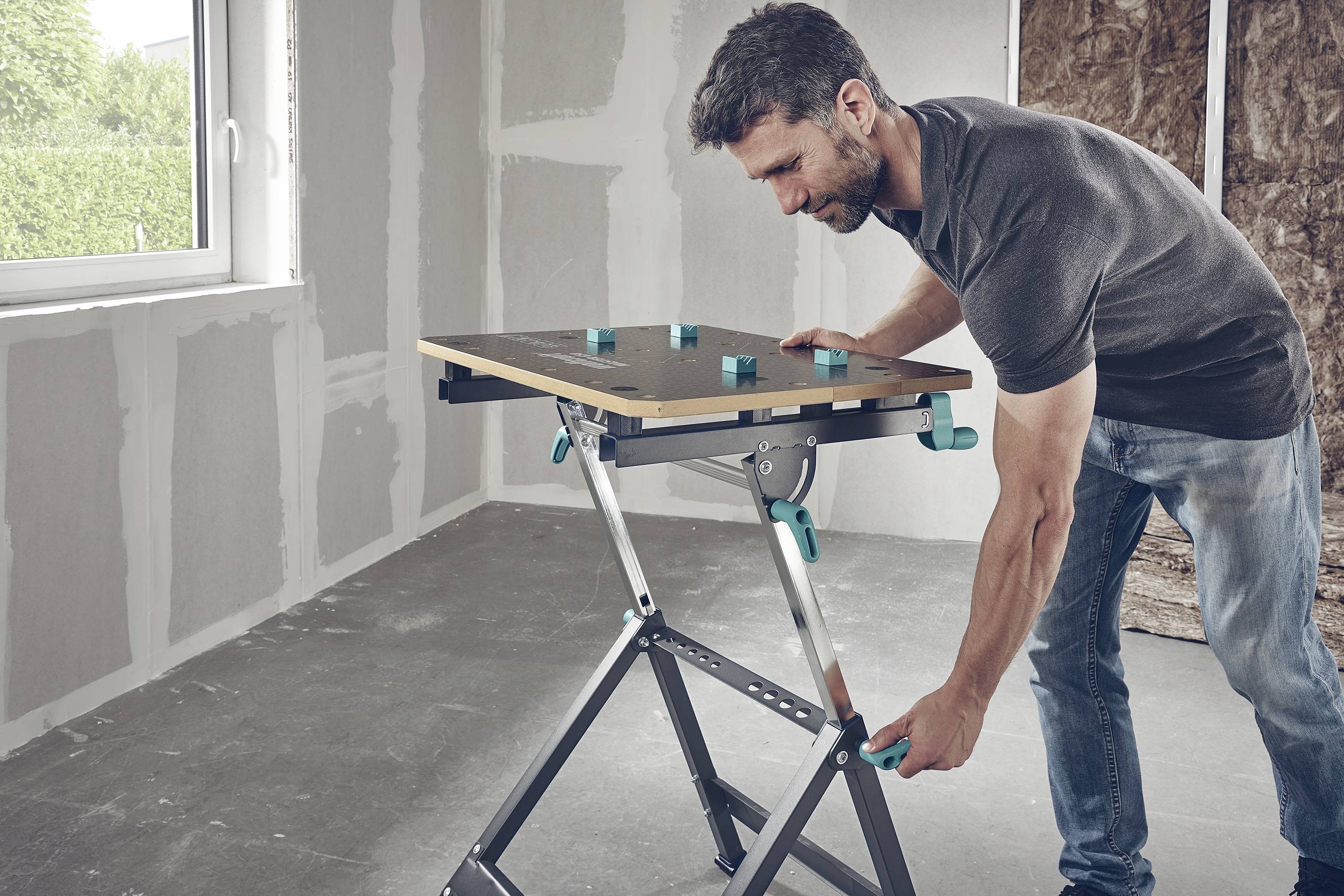 Mężczyzna stoi w niedokończonym pomieszczeniu i reguluje wysokość przenośnego stołu roboczego.