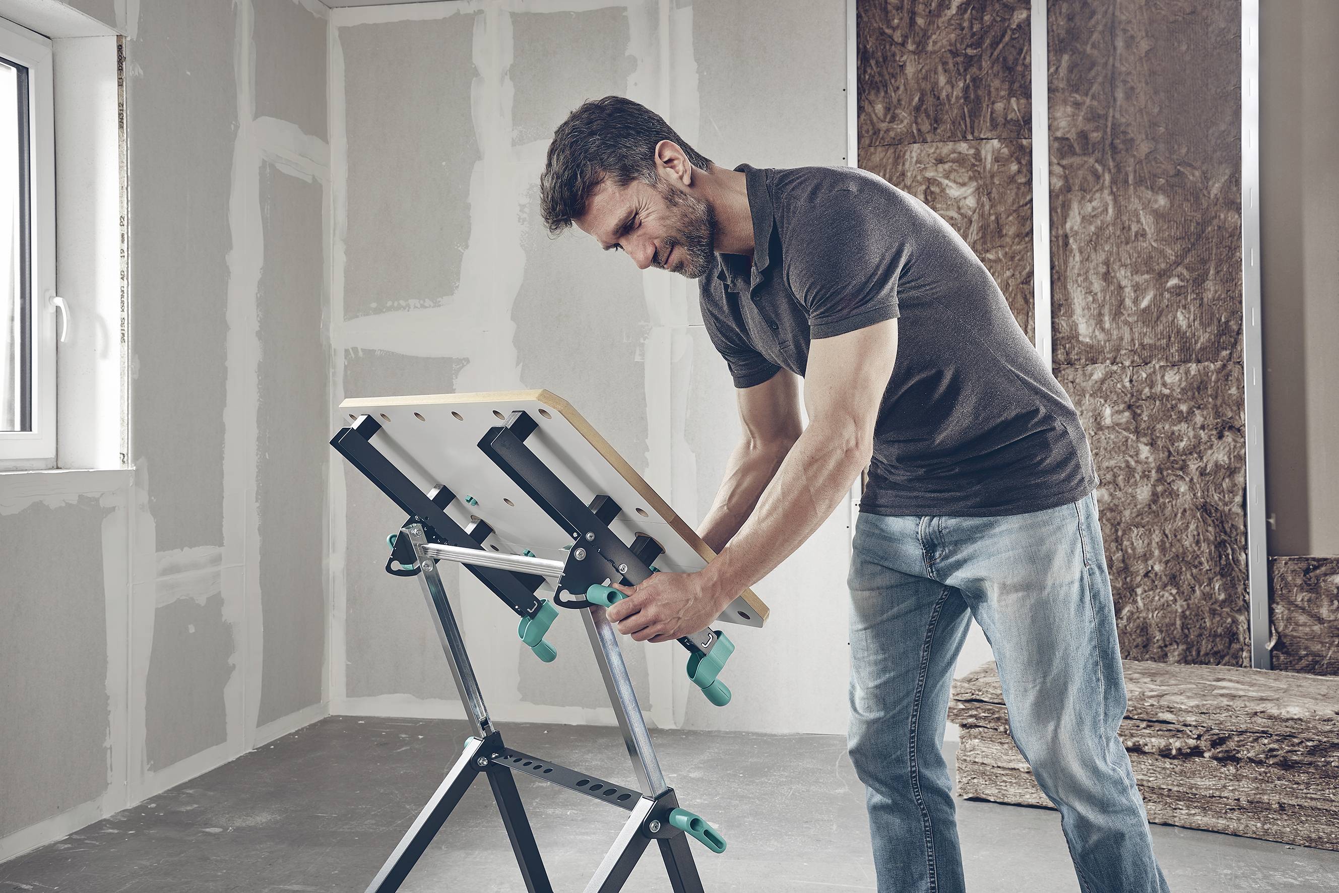Mężczyzna w surowym pomieszczeniu reguluje stół roboczy ze zielonymi zaciskami. Światło pada na niego przez okno.