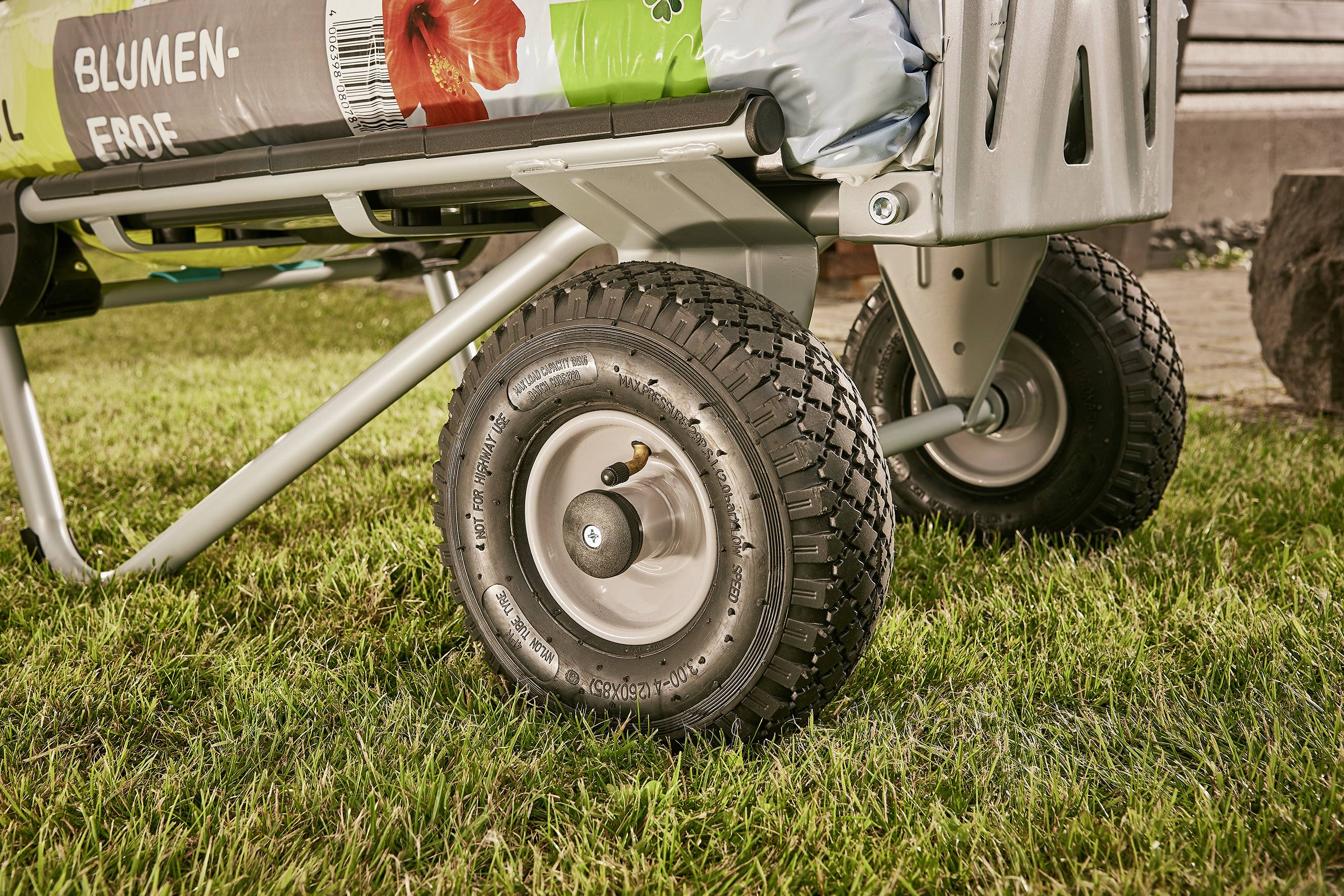 Zbliżenie wózka metalowego z dużymi kołami na łące, załadowanego workami ziemi kwiatowej z nadrukiem czerwonego kwiatu.