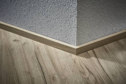 Róg pomieszczenia z drewnianą podłogą i szarą, strukturalną ścianą. Drewniana listwa przypodłogowa łączy podłogę ze ścianą w harmonijny sposób.