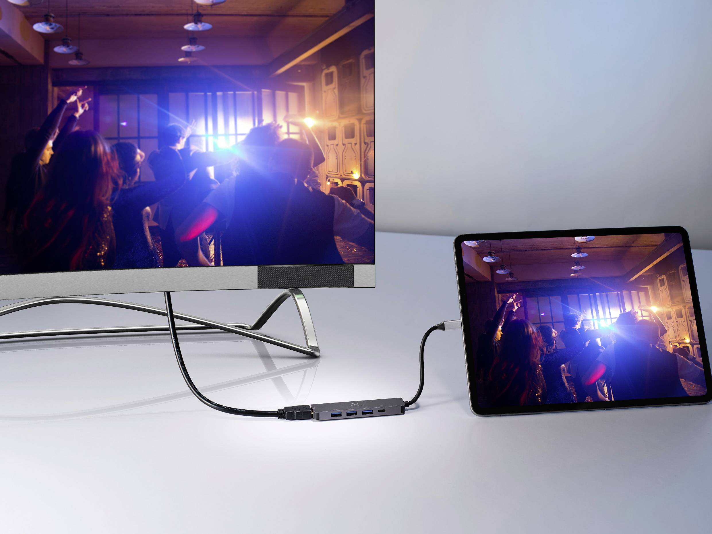 Tablet jest podłączony przez adapter do monitora, który wyświetla ten sam obraz z imprezy. Scena przedstawia ludzi bawiących się na przyjęciu.