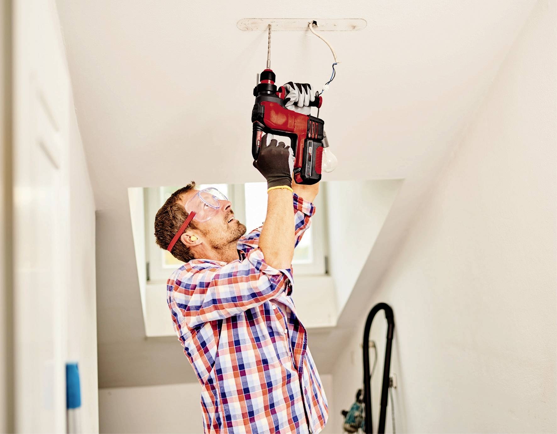 Mężczyzna w kratowanej koszuli i okularach ochronnych używa wiertarki, aby wywiercić otwór w suficie. W tle stoi drabina.