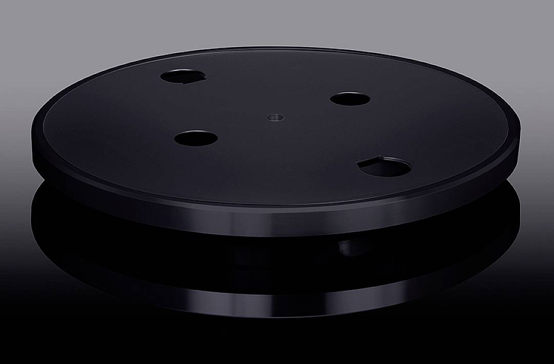 Czarna okrągła tarcza z kilkoma strategicznie rozmieszczonymi wgłębieniami, spoczywająca na odblaskowej powierzchni.