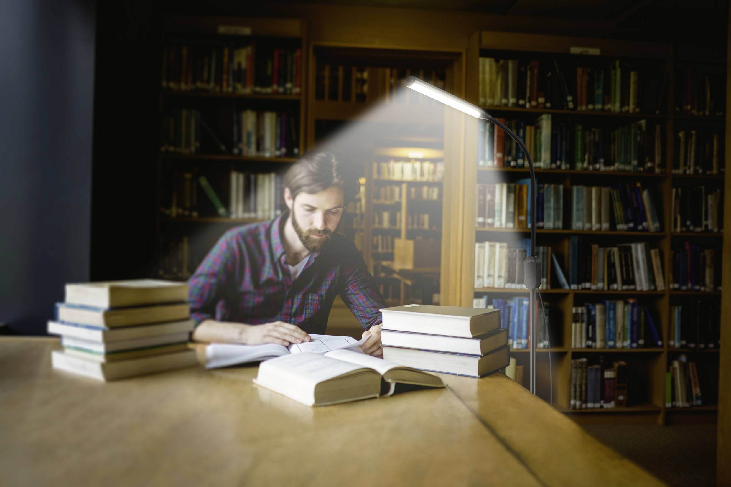 Osoba czyta w bibliotece, otoczona stosami książek. Lampa oświetla stół.