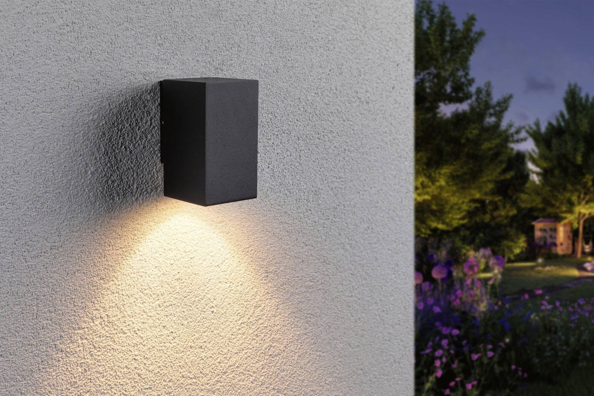 Czarna lampa ścienna świecąca w dół na szarej ścianie, obok ogród o zmierzchu.