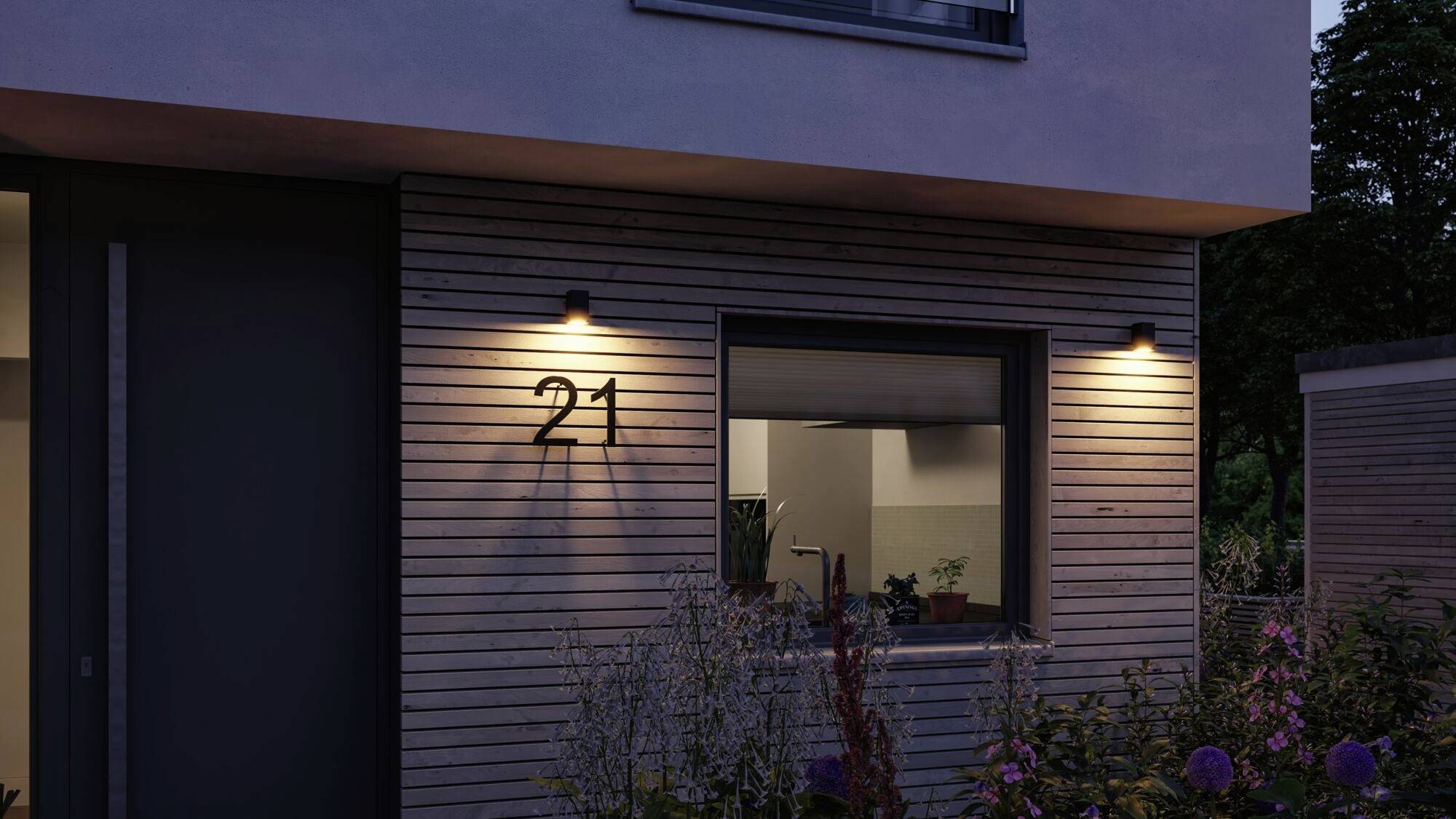 Nowoczesny dom nocą, oświetlony przez dwie lampy zewnętrzne. Obok okna widoczny jest numer domu '21'.