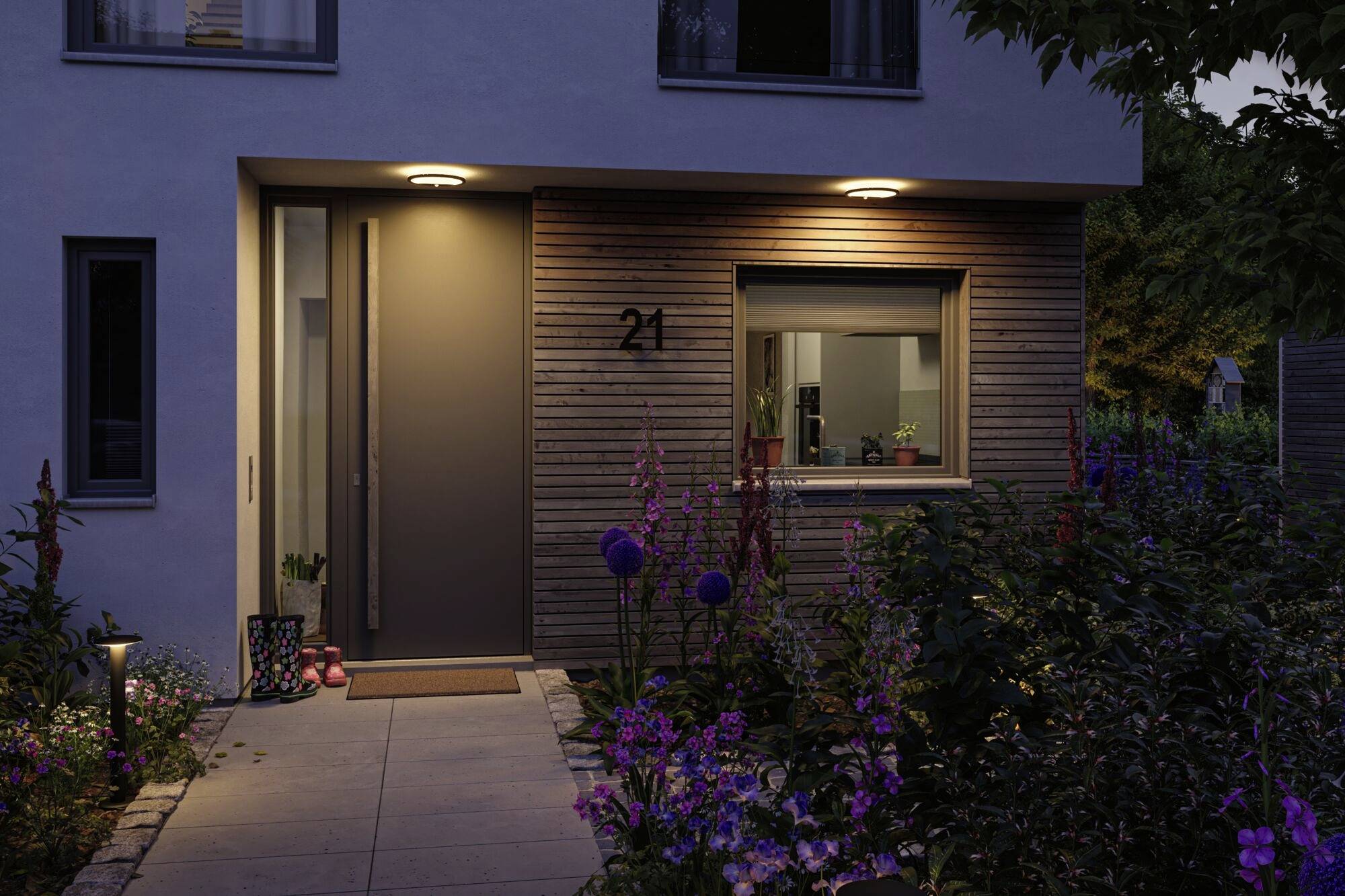 Wejście do nowoczesnego domu wieczorem, oświetlone, z roślinami i kwiatami wzdłuż ścieżki, numer domu '21' obok drzwi.