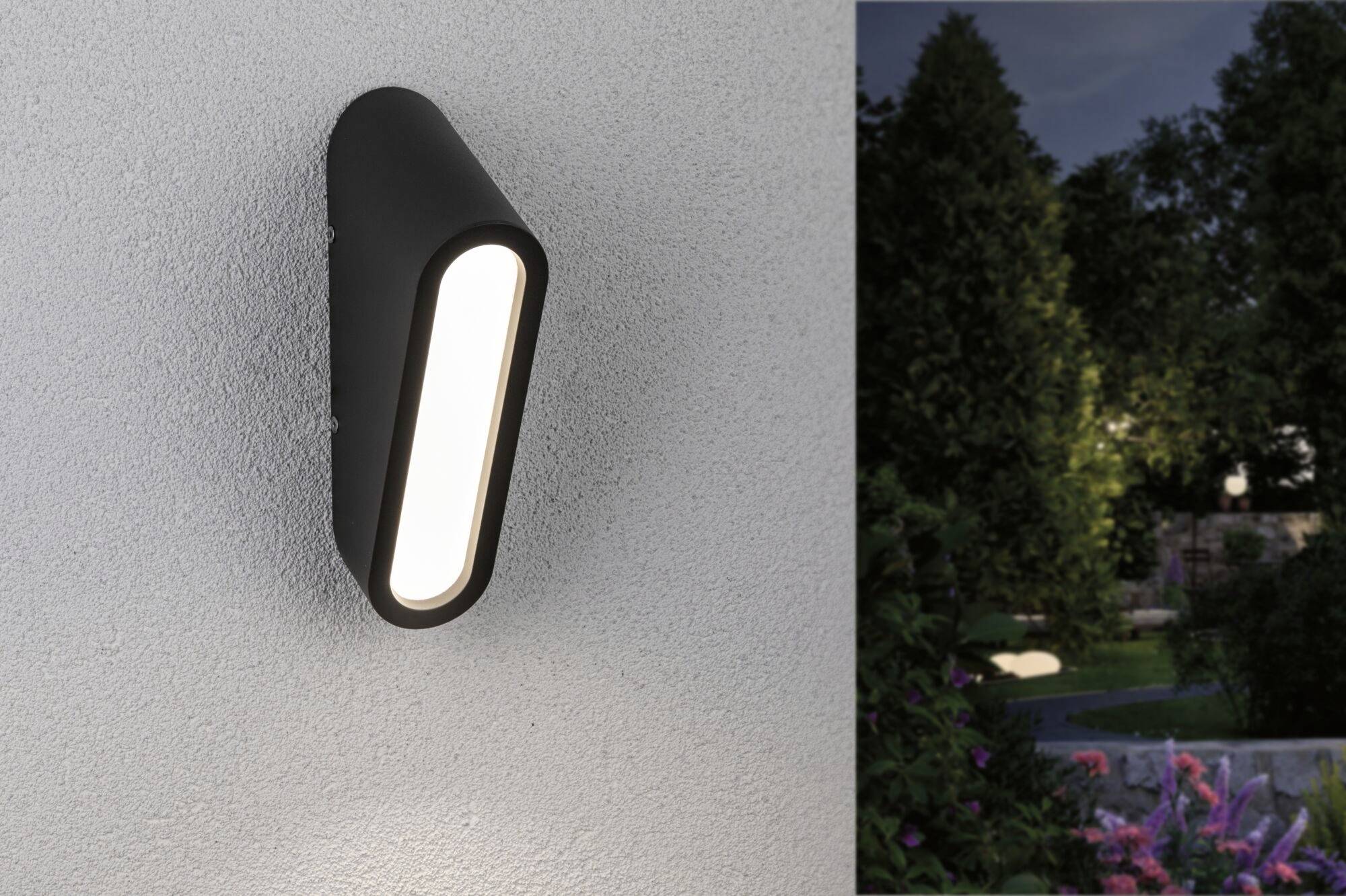 Czarna lampa zewnętrzna świeci na ścianie, oświetlając fragment ogrodu. W ogrodzie widoczne są rośliny i oświetlona ścieżka.